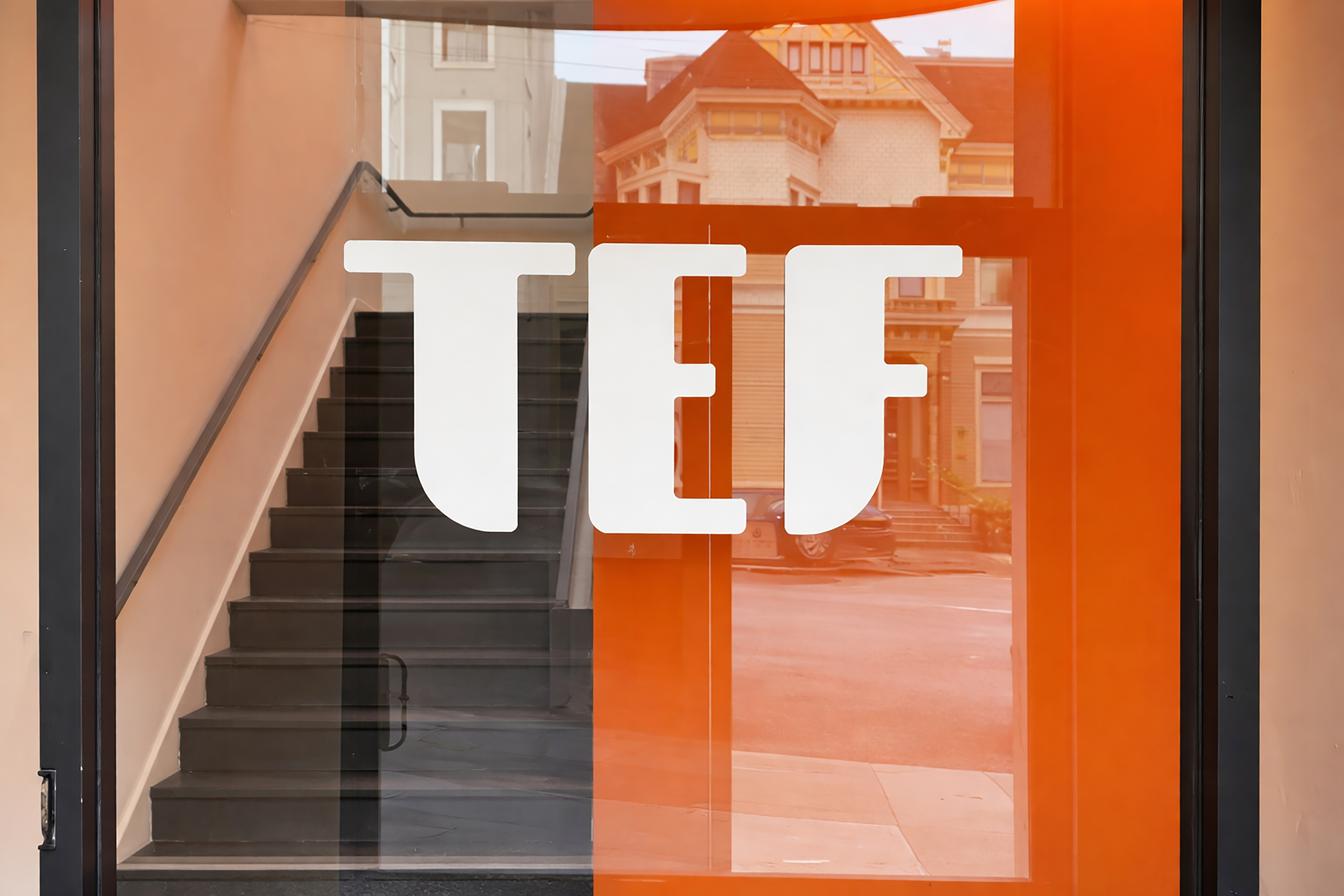 Close-up view of TEF’s office entrance featuring the white “TEF” logo on an orange-tinted glass door, with softened reflections of nearby Victorian-style San Francisco houses visible beyond.