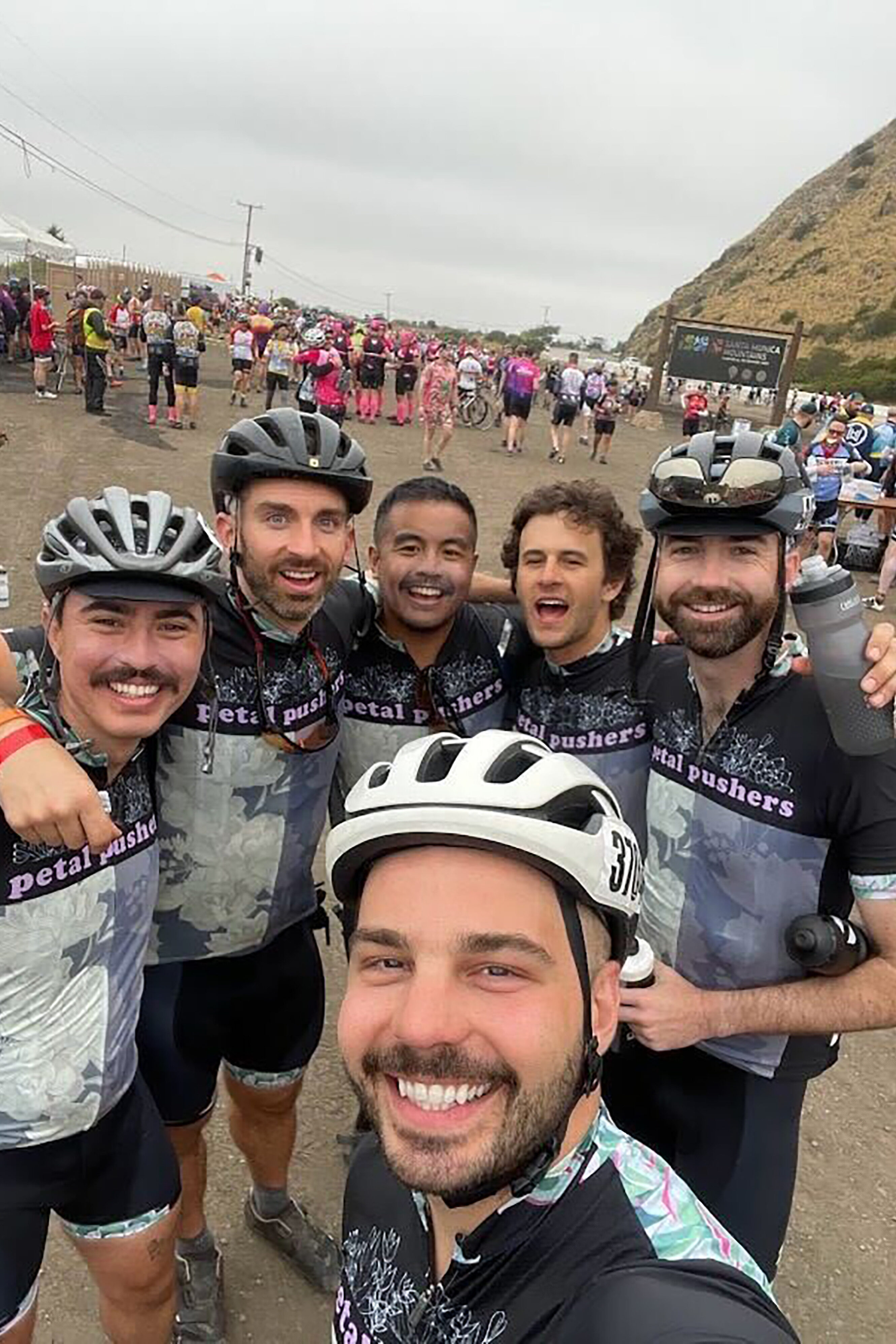 OJ with group of cyclists in matching jerseys pose together smiling at an outdoor cycling event with hills and other riders in the background.