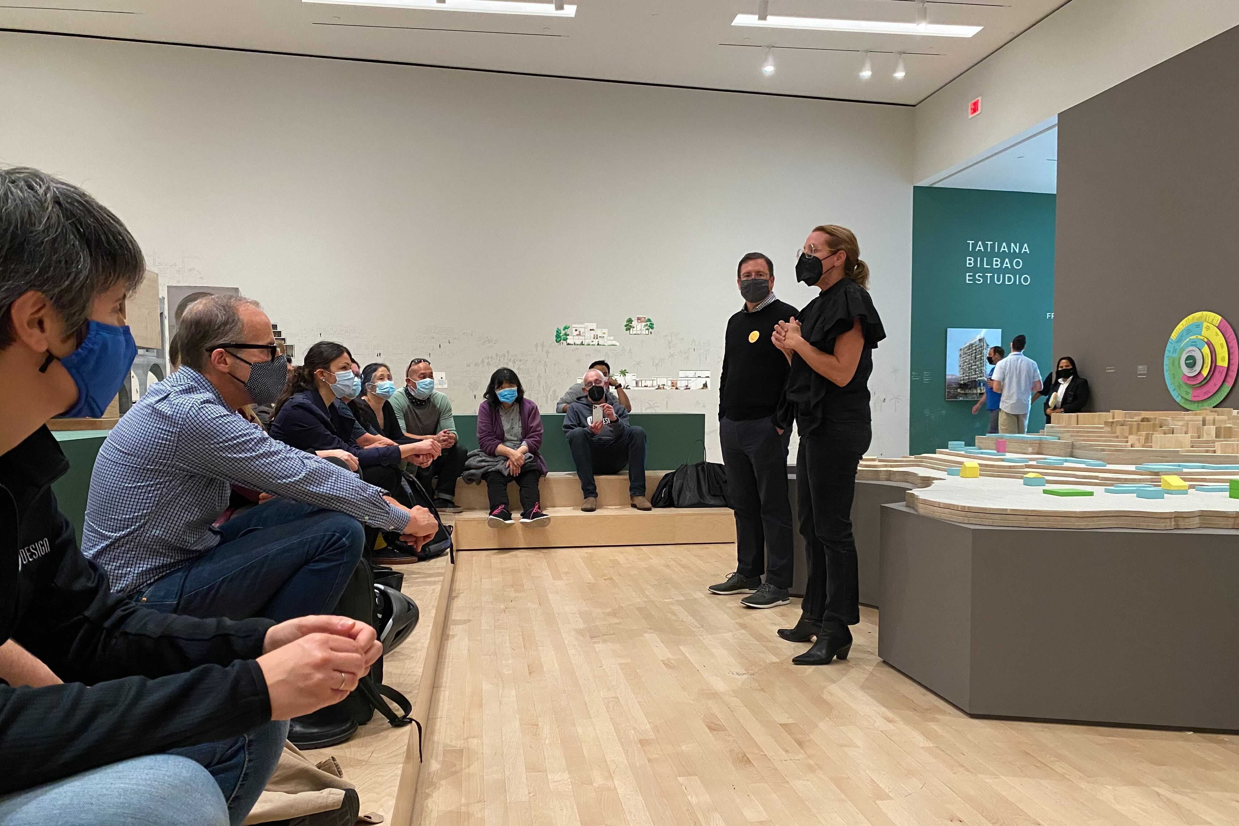Group of masked attendees listening to a presentation beside architectural models in a museum gallery space.