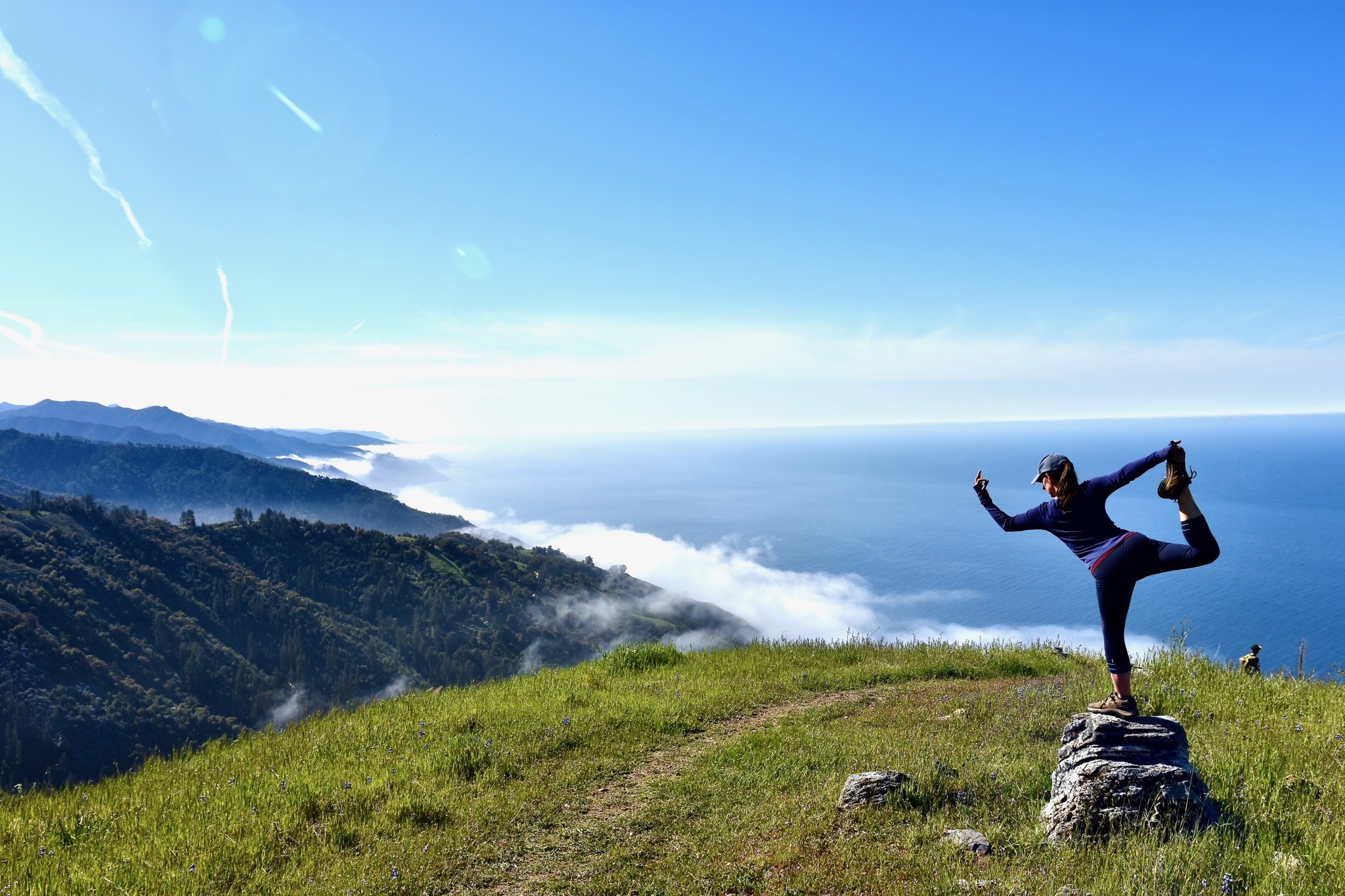 A person in hiking gear in a yoga pose on a green hill overlooking a wide bay below