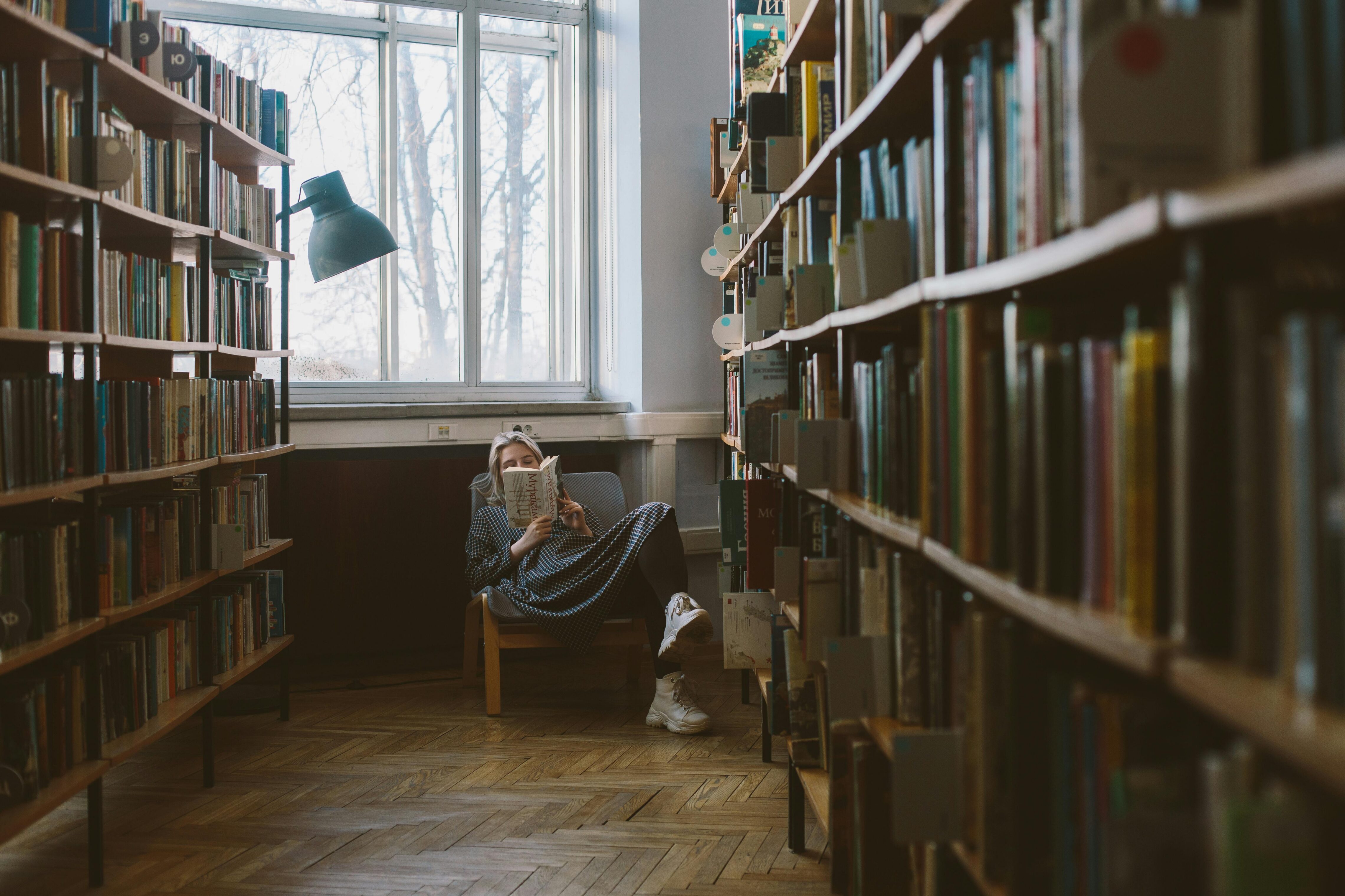 A woman reading a book at the end of an aisle of full bookshelves