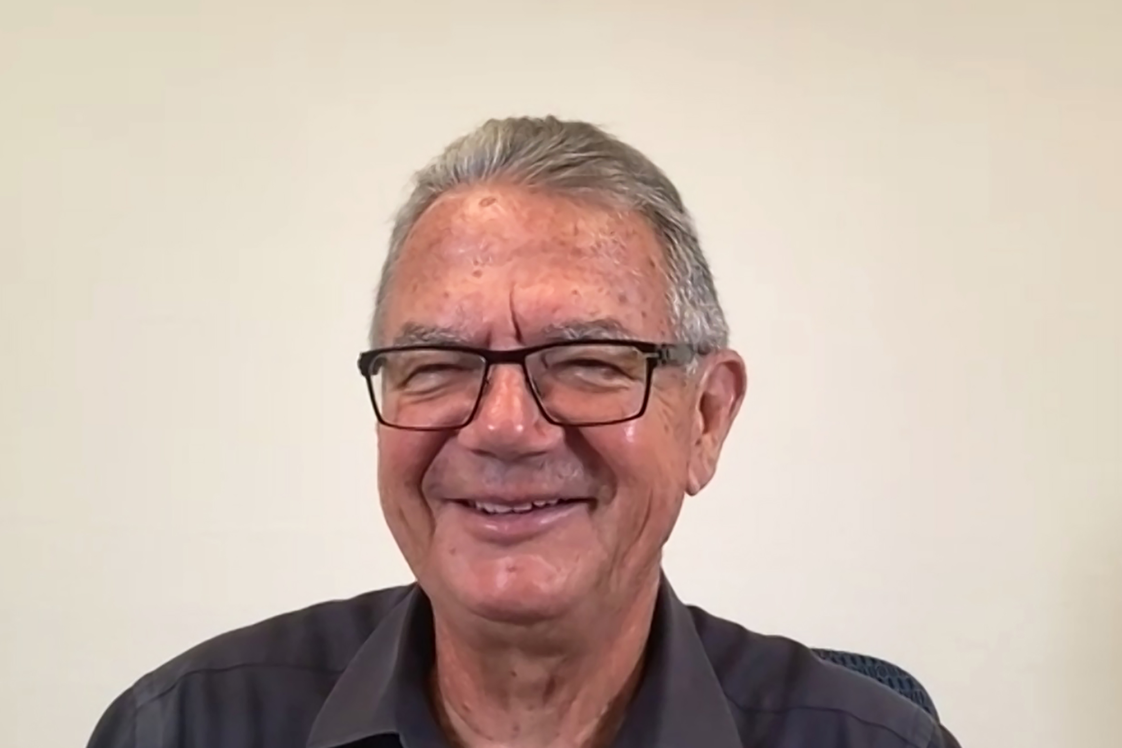 Patrick Bell smiling with short gray hair and glasses, wearing a dark button-up shirt, photographed against a plain light-colored background.