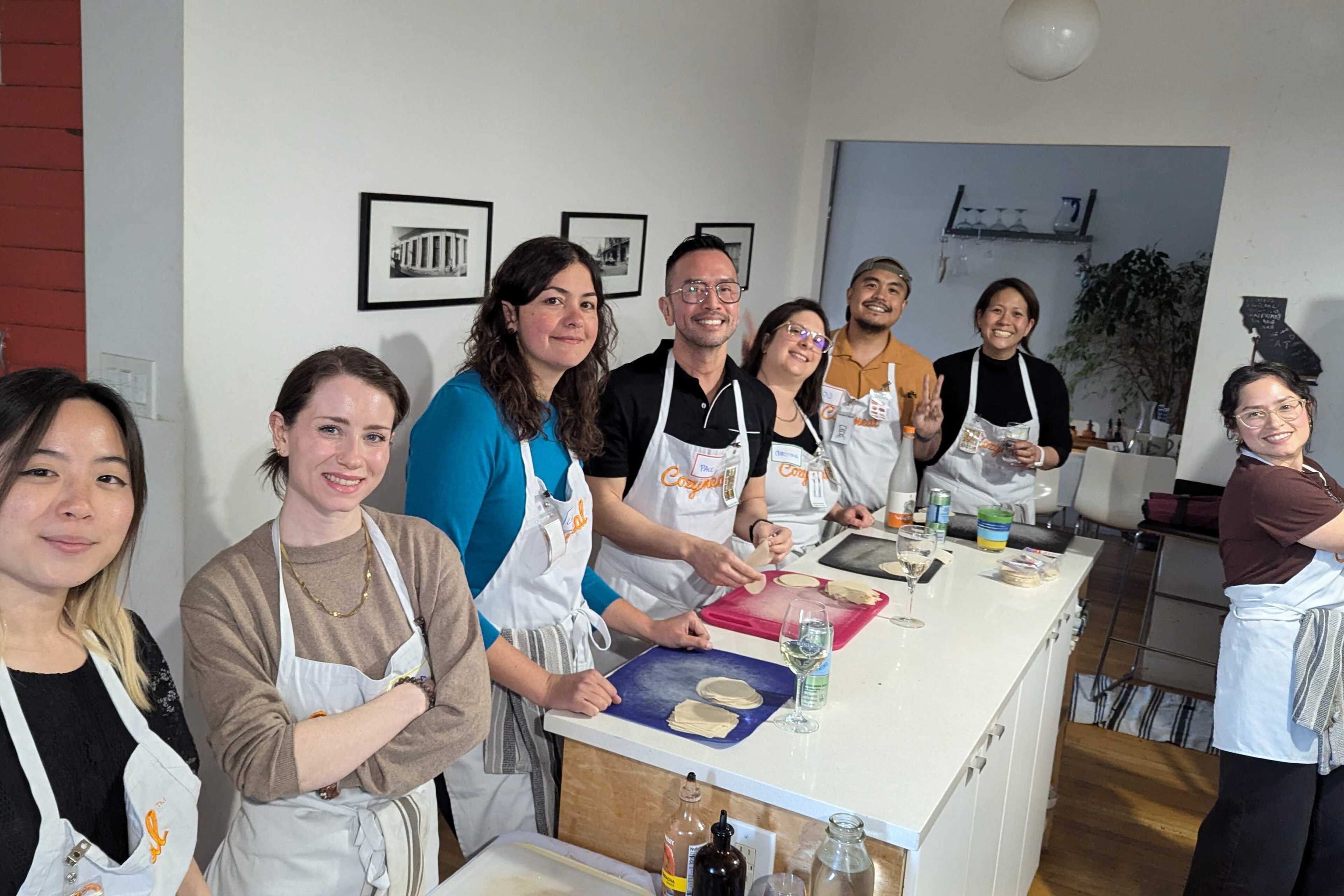 A group of coworkers wearing aprons stand around a kitchen island, smiling while preparing food together during a team cooking activity.