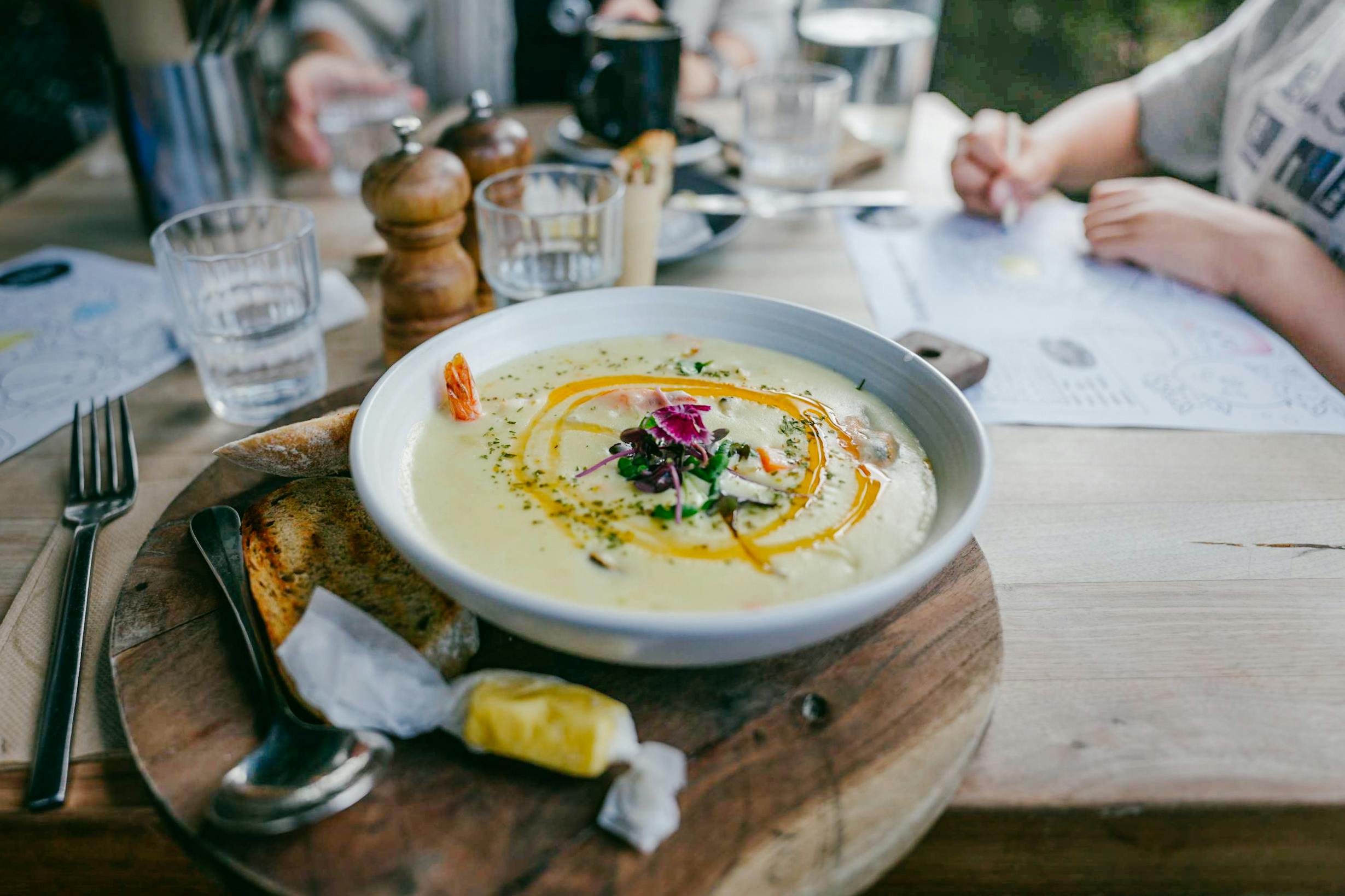 A bowl of pale yellow soup with oil and garnishes set on a table with people around.