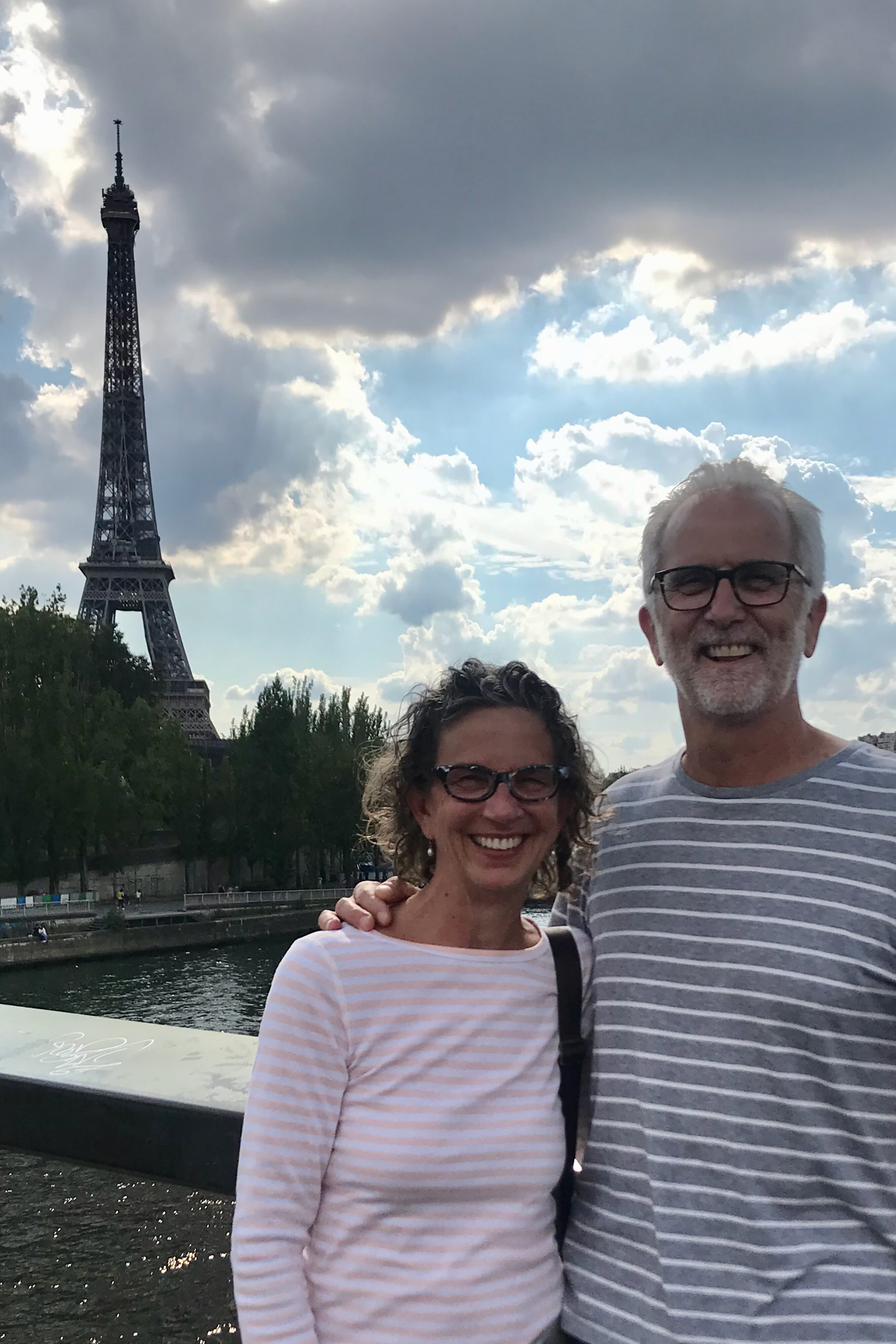 Two people smile while standing by a river with the Eiffel Tower visible in the background under a partly cloudy sky.