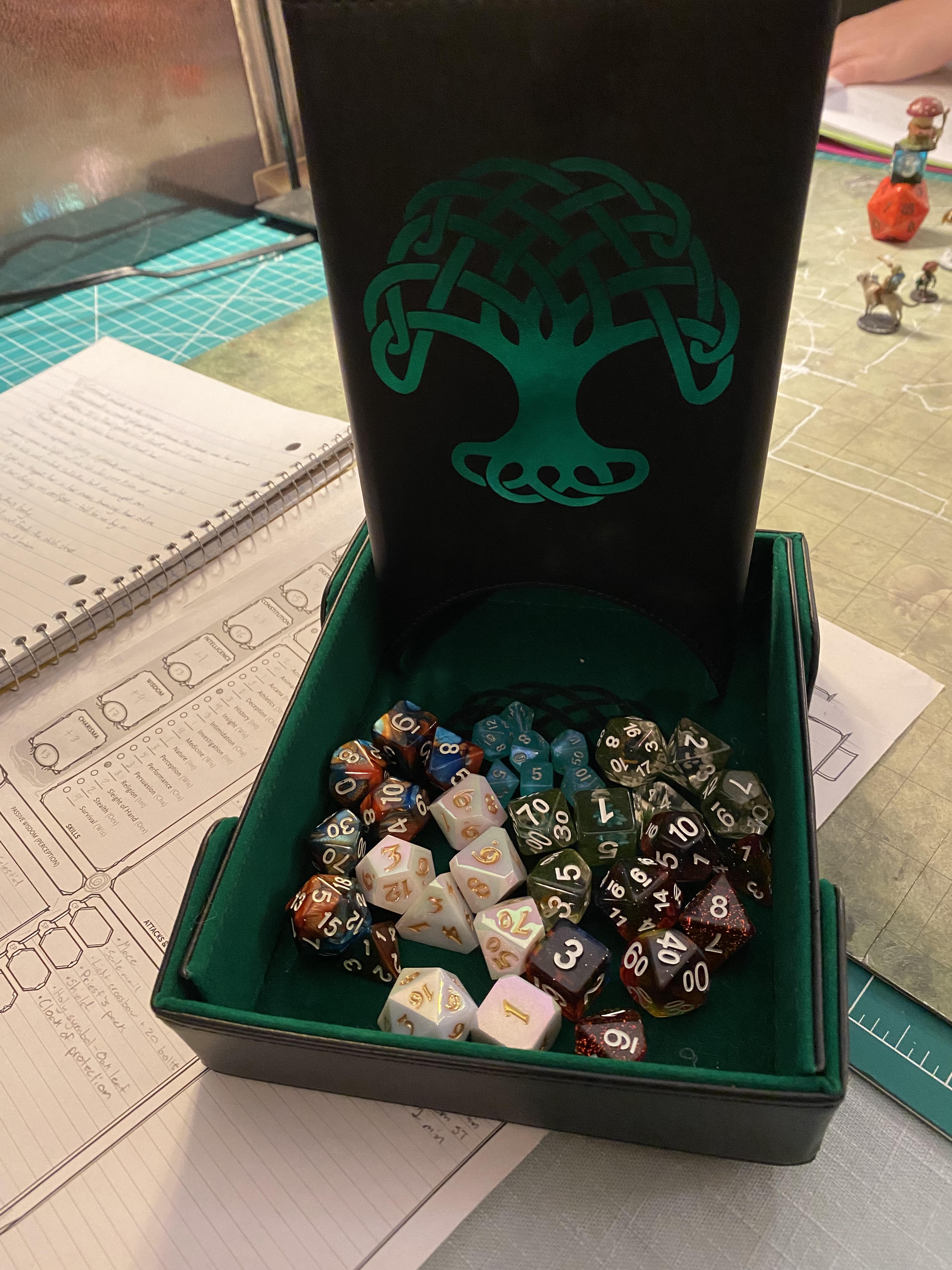 Dice roller tower with green celtic looking tree on in filled with several different colored dice on top of a Dungeons and Dragons player sheet.