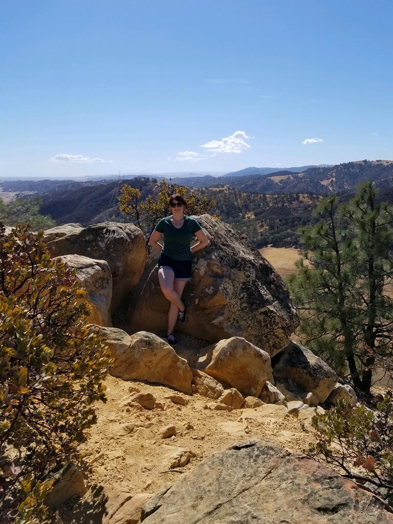 A woman in hiking clothes stands among large rocks on a sunlit mountain overlook, with dry hills, scattered trees, and a clear blue sky in the distance.