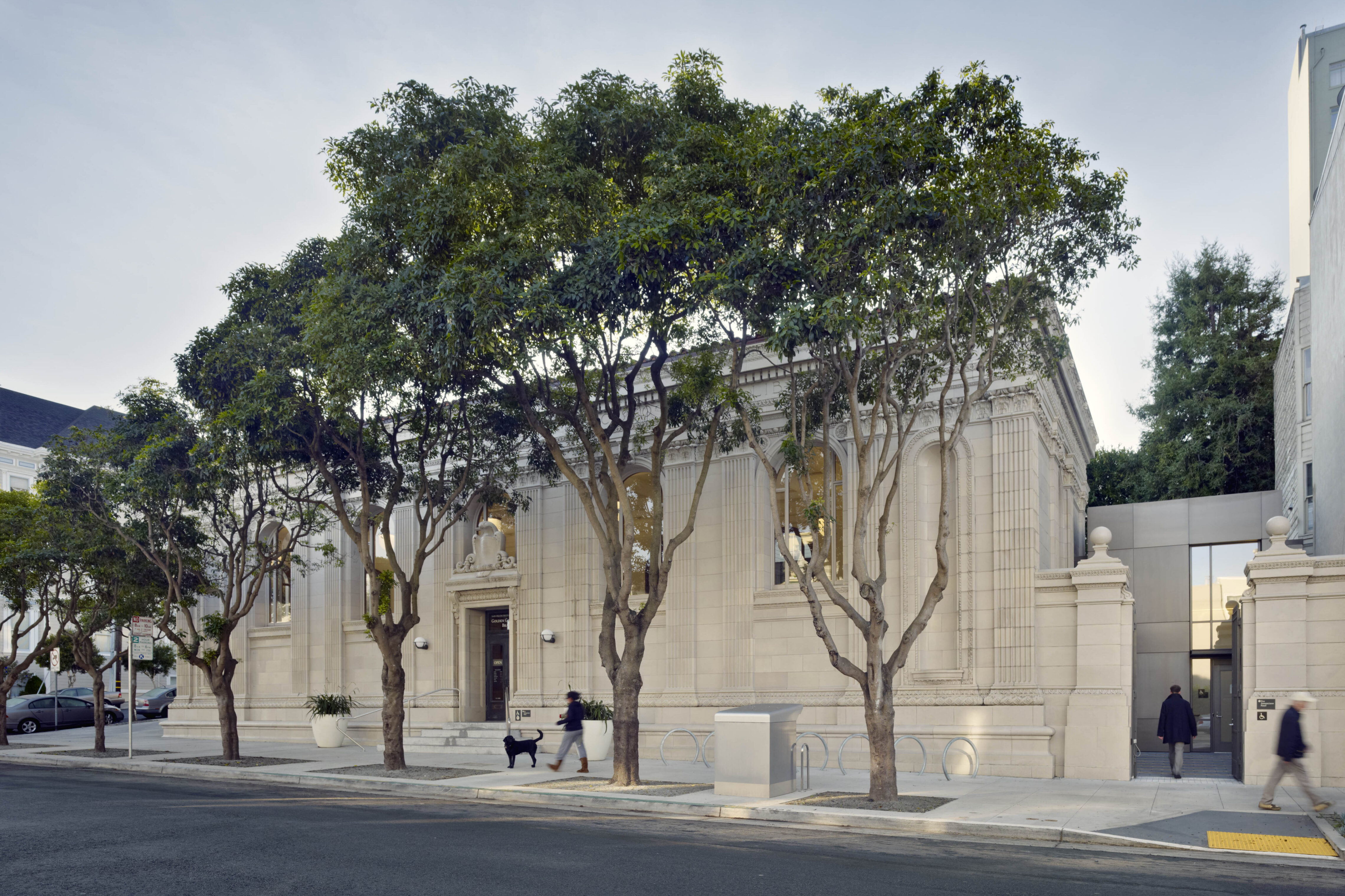 Historic library building with ornate stone façade and street trees, connected to a contemporary addition along a quiet sidewalk.