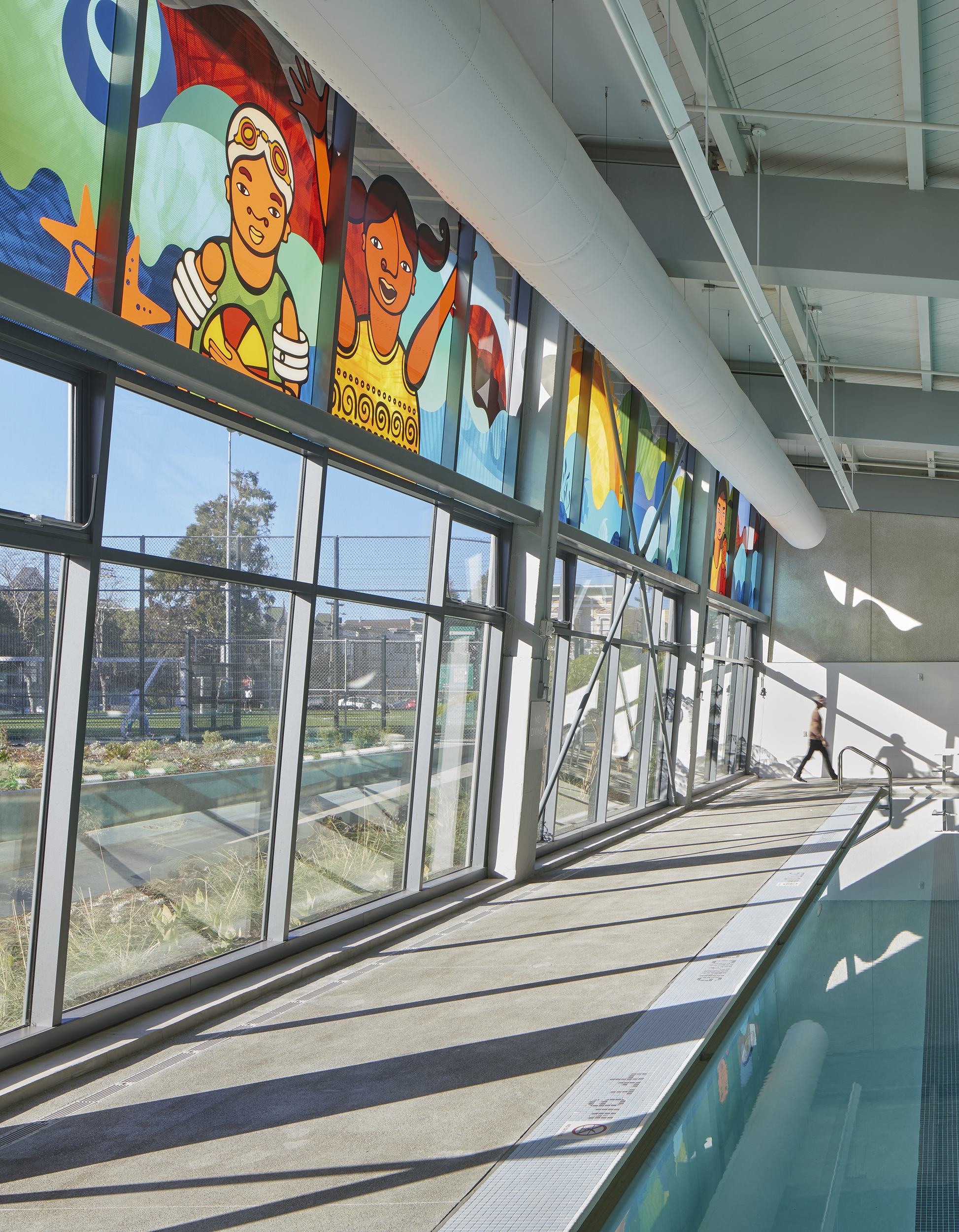 Colorful glass art panels above an indoor pool, bringing light, movement, and community-focused imagery into the aquatic facility.