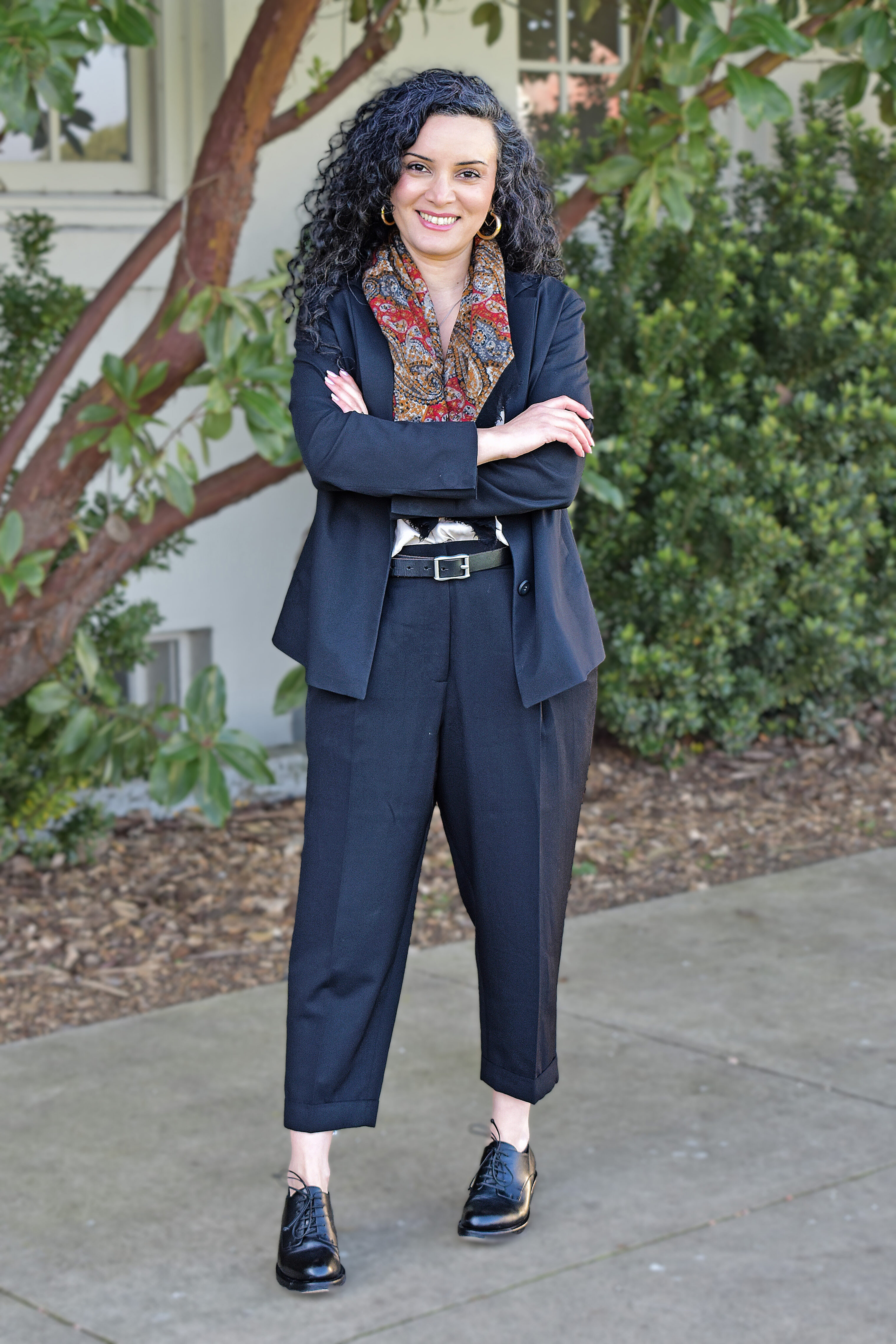 Maryam standing outdoors on a sidewalk with arms crossed, smiling toward the camera, wearing a dark suit with a patterned scarf and dress shoes, with greenery and a building façade in the background.