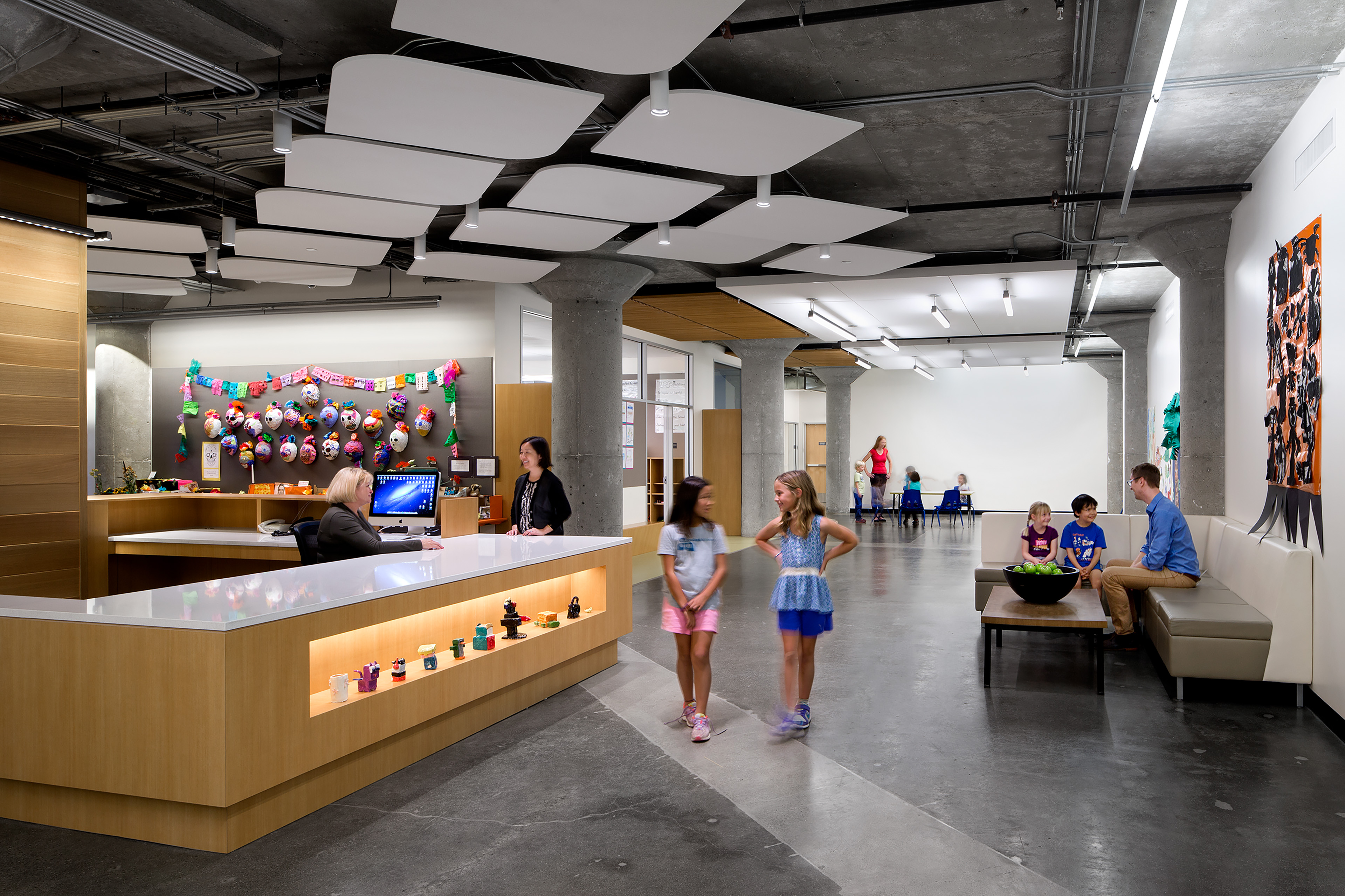 Students walk through a bright school lobby with a reception desk, exposed concrete columns, and suspended acoustic ceiling panels.