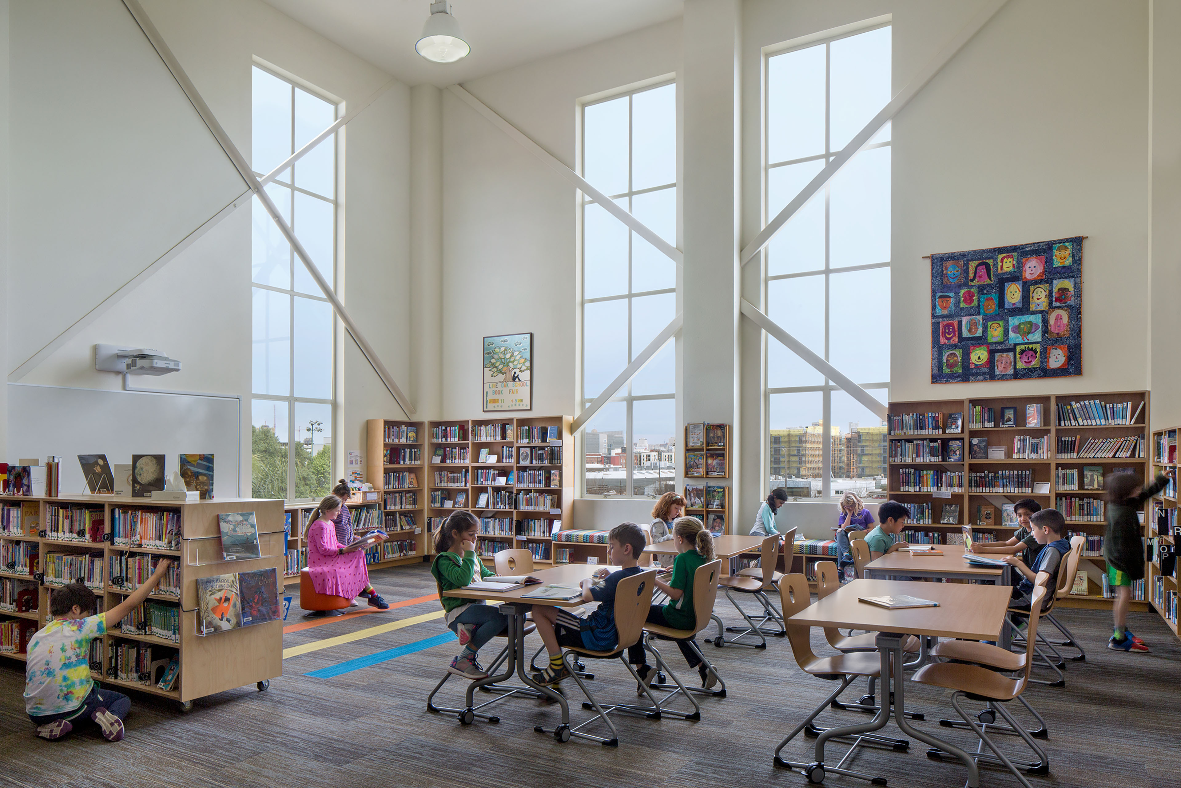 Students read and study at tables in a library filled with natural light from tall windows.
