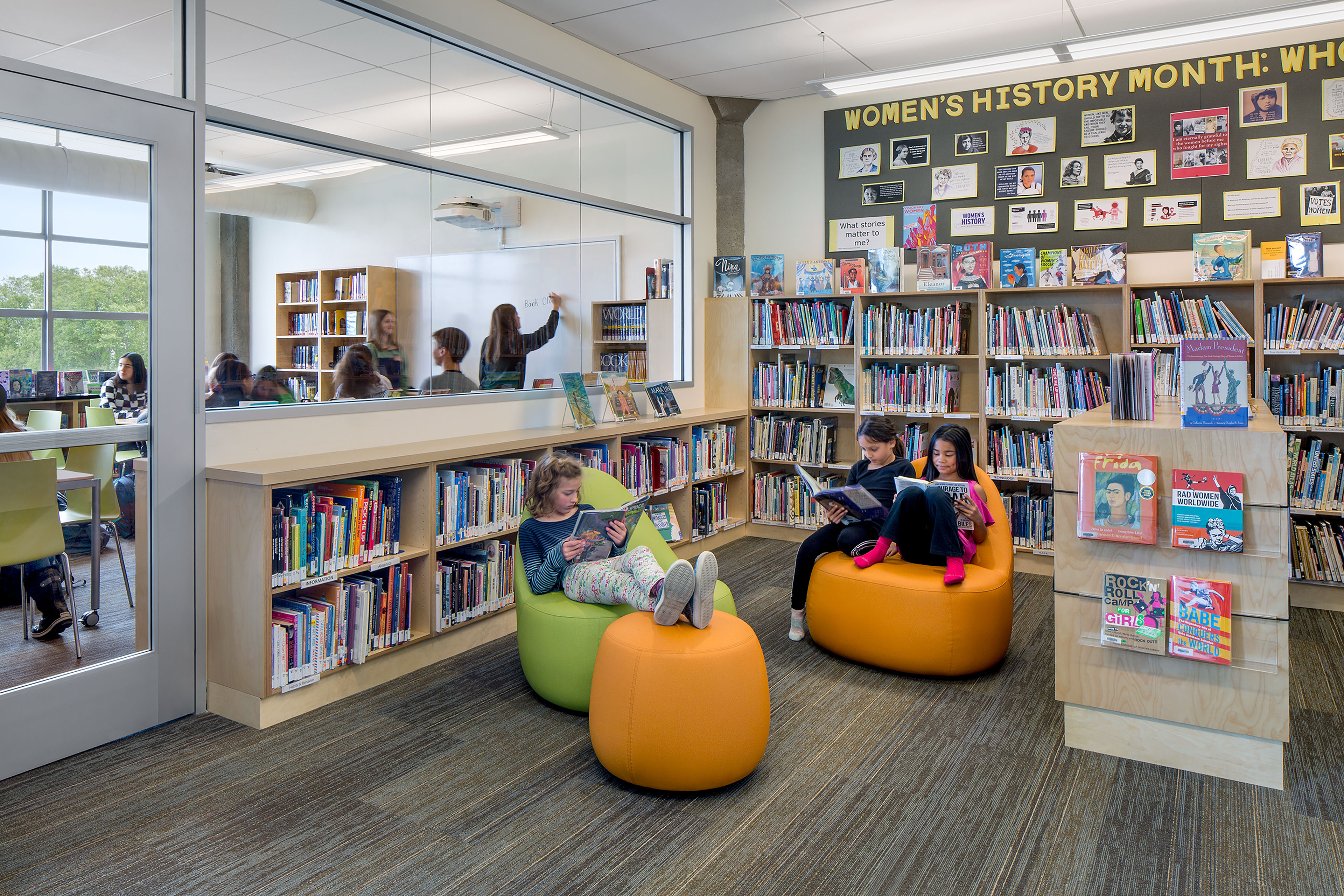 School library with floor-to-ceiling bookshelves and a Women’s History Month display. Three children read on colorful lounge chairs and ottomans, while a glass wall reveals a classroom lesson in progress next door.