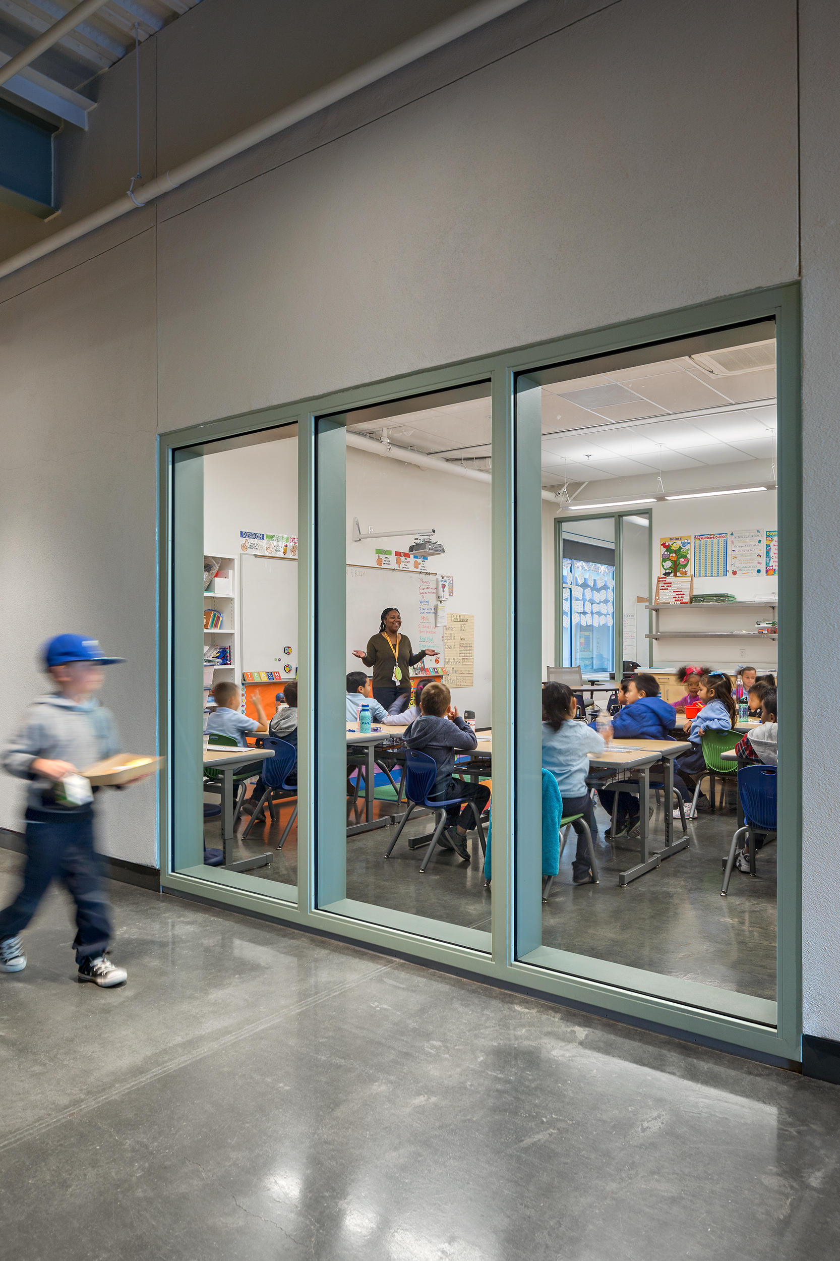 Teacher leads a lesson while students sit at tables inside a classroom, viewed through a large interior glass window from the hallway.