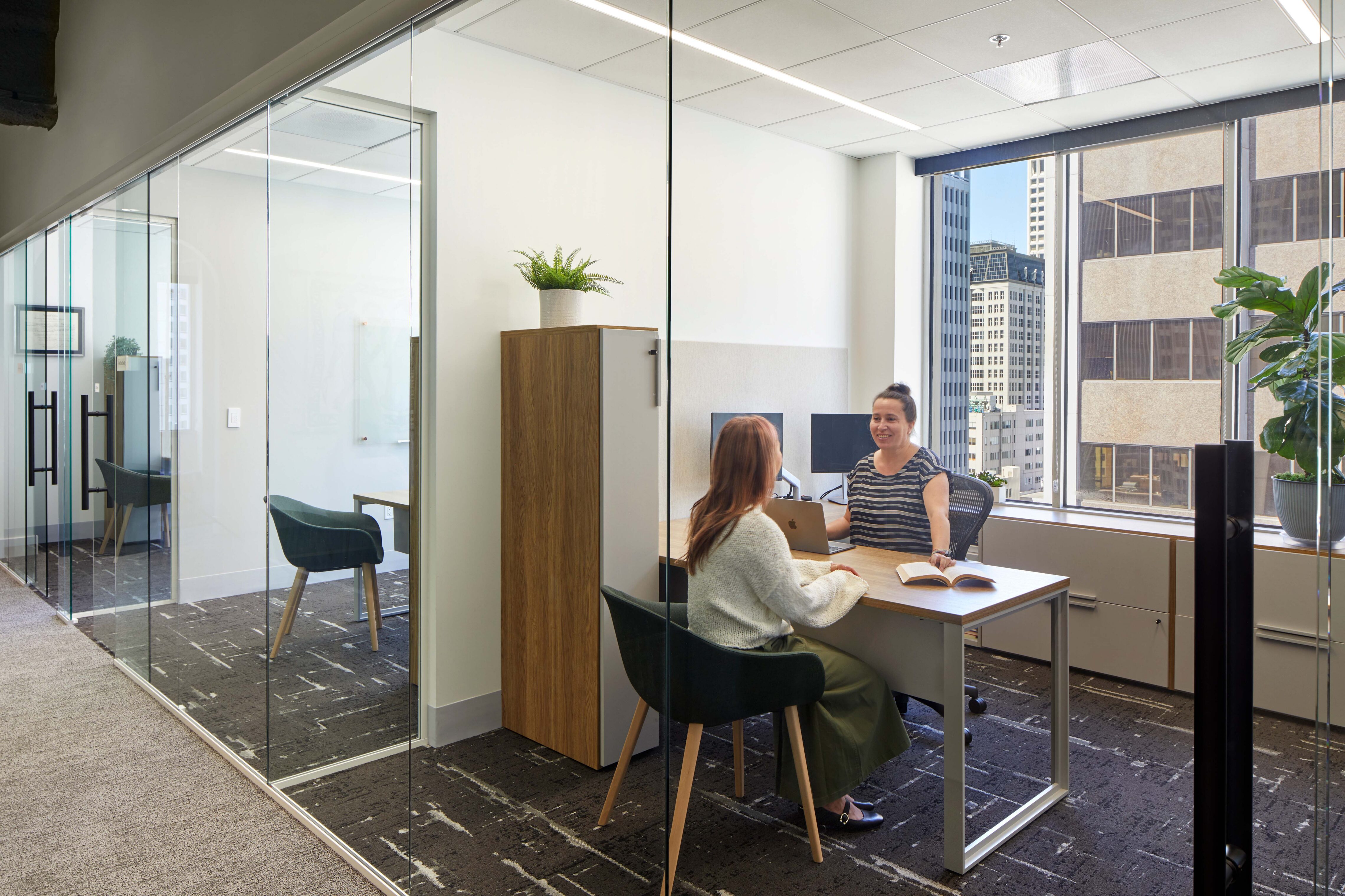 Two colleagues meeting at a desk in a glass-walled office with views of nearby downtown buildings.