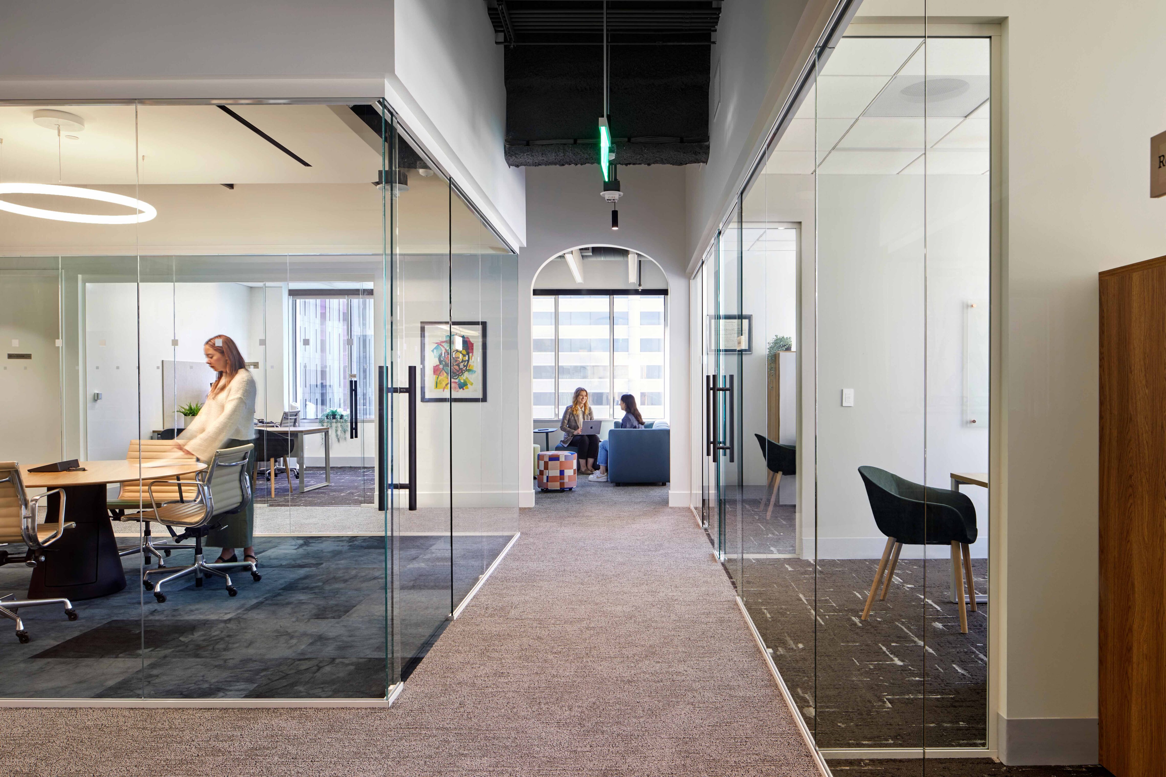 Office corridor lined with glass meeting rooms leading to a casual seating area where two people work together.