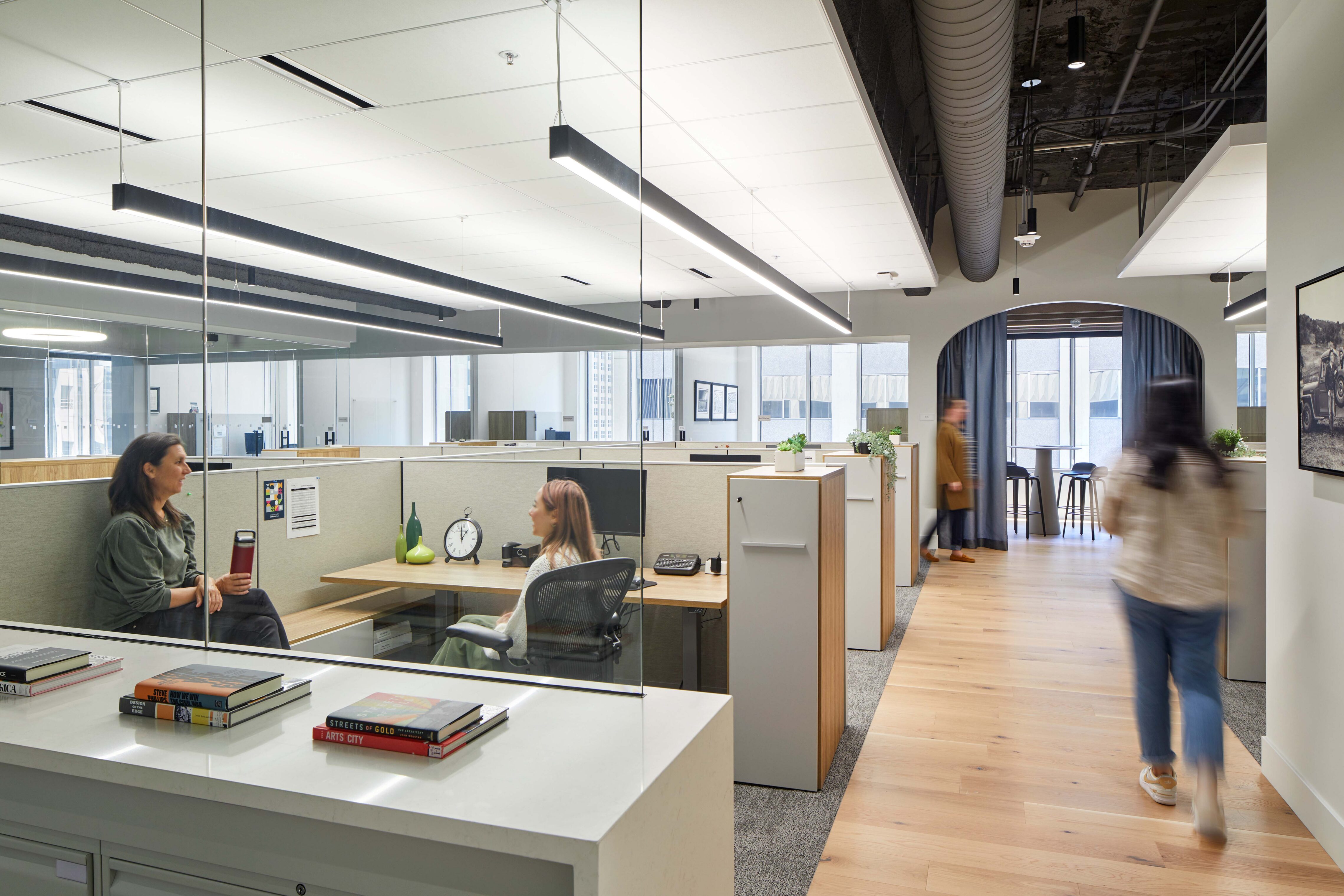 Open office area with cubicles and glass partitions where colleagues talk at a workstation while others move through the space.
