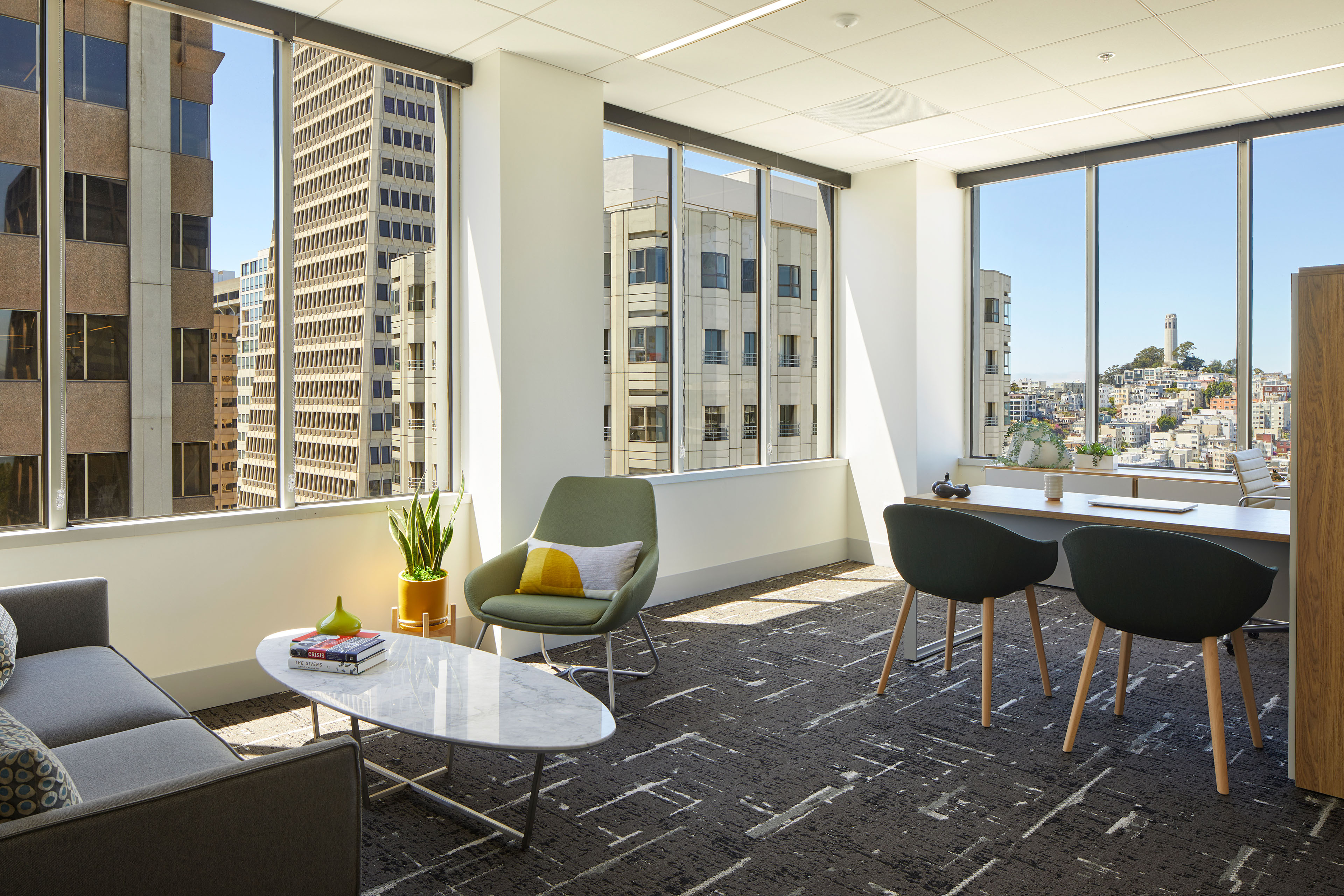 Sunlit private office and meeting space with floor-to-ceiling windows overlooking downtown San Francisco and Coit Tower. A gray sofa, green lounge chair, marble coffee table, and dark upholstered meeting chairs sit on patterned carpet, with a wood desk positioned beside the corner windows.