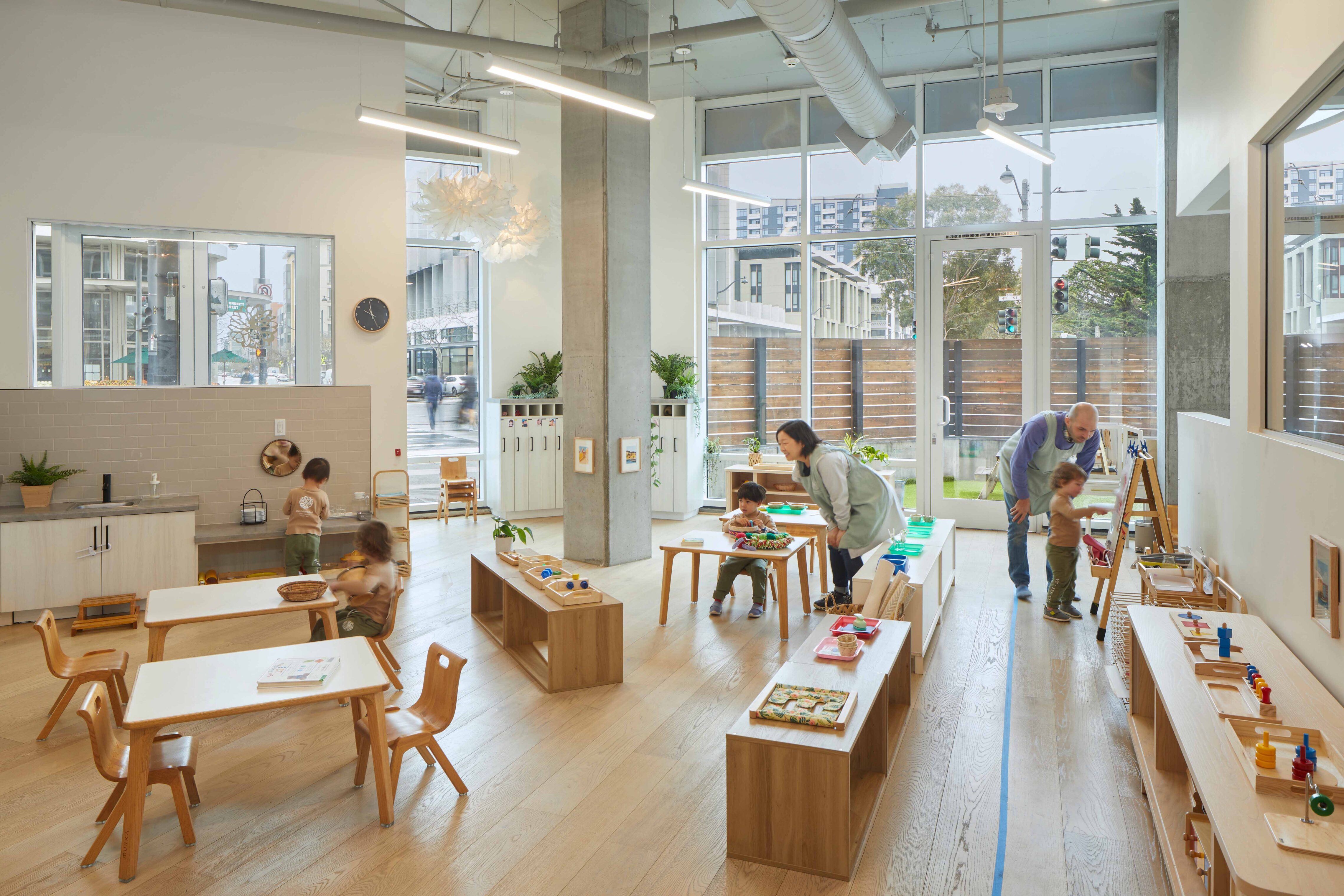 Teachers assisting children with hands-on activities in a spacious preschool classroom with natural light, wood furnishings, and organized learning materials.