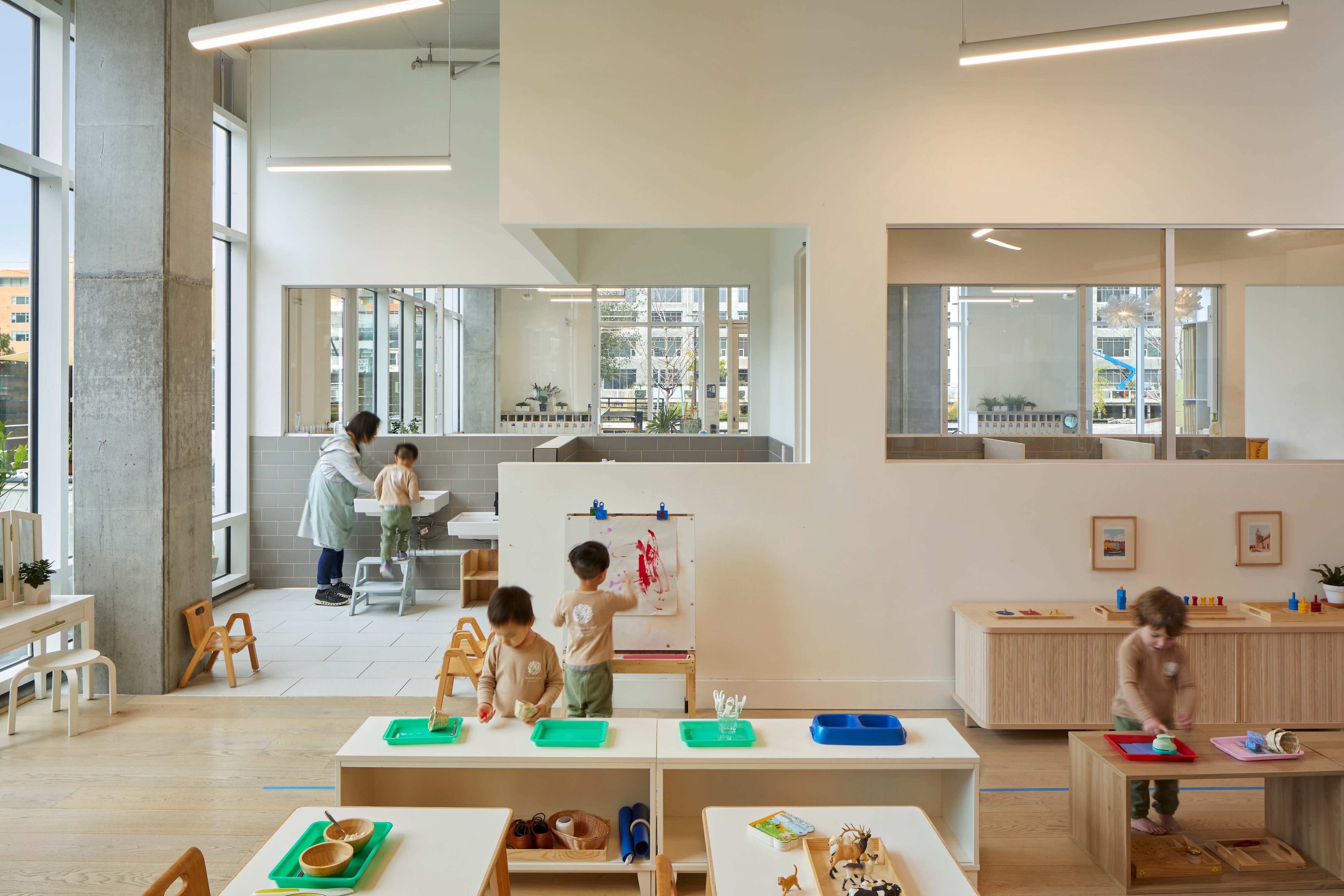 Young children working at low tables and activity stations in a preschool classroom with a sink area and interior viewing windows.