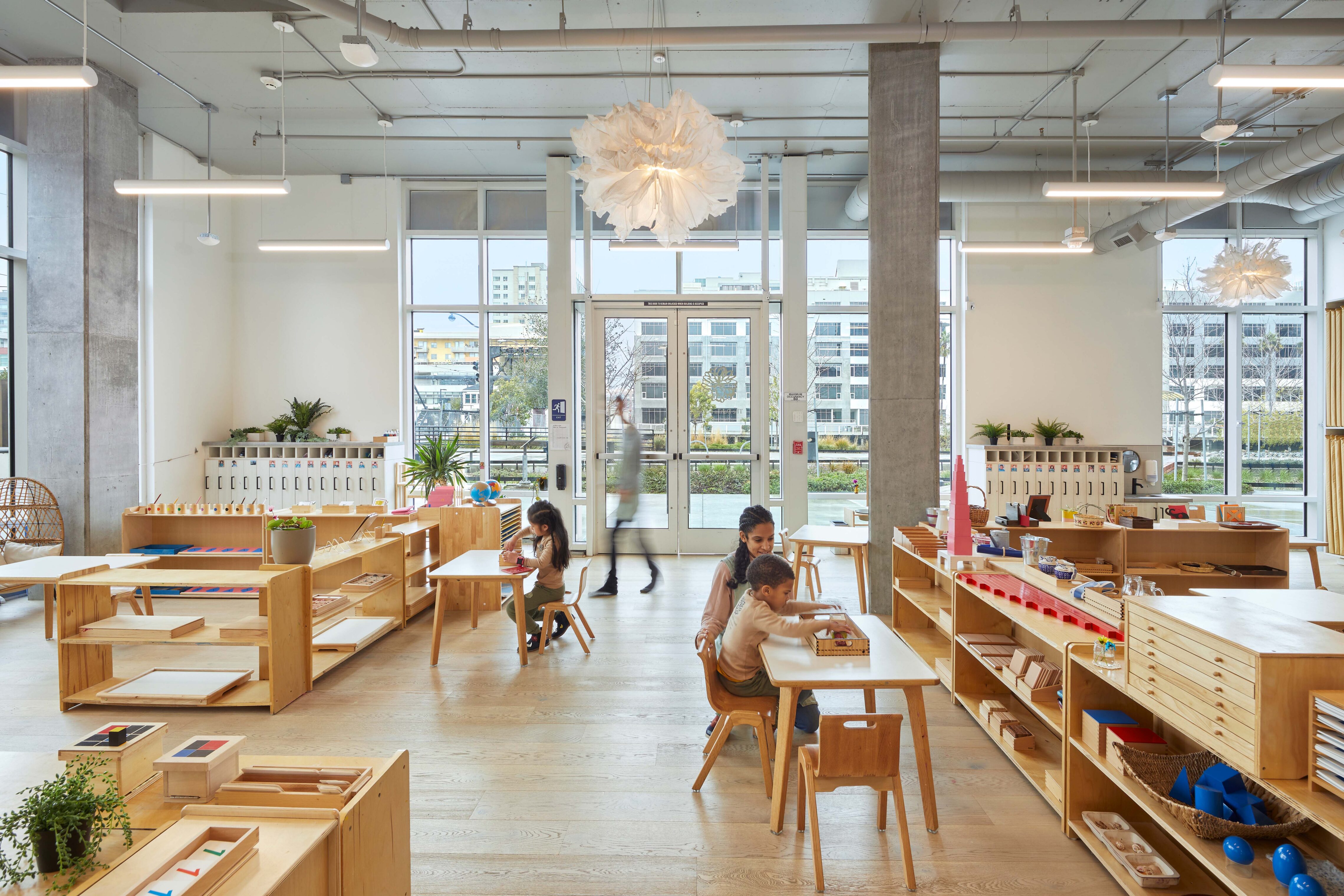 Children and a teacher engaged in activities at small tables in a bright classroom with wooden shelving, tall windows, and pendant lighting.