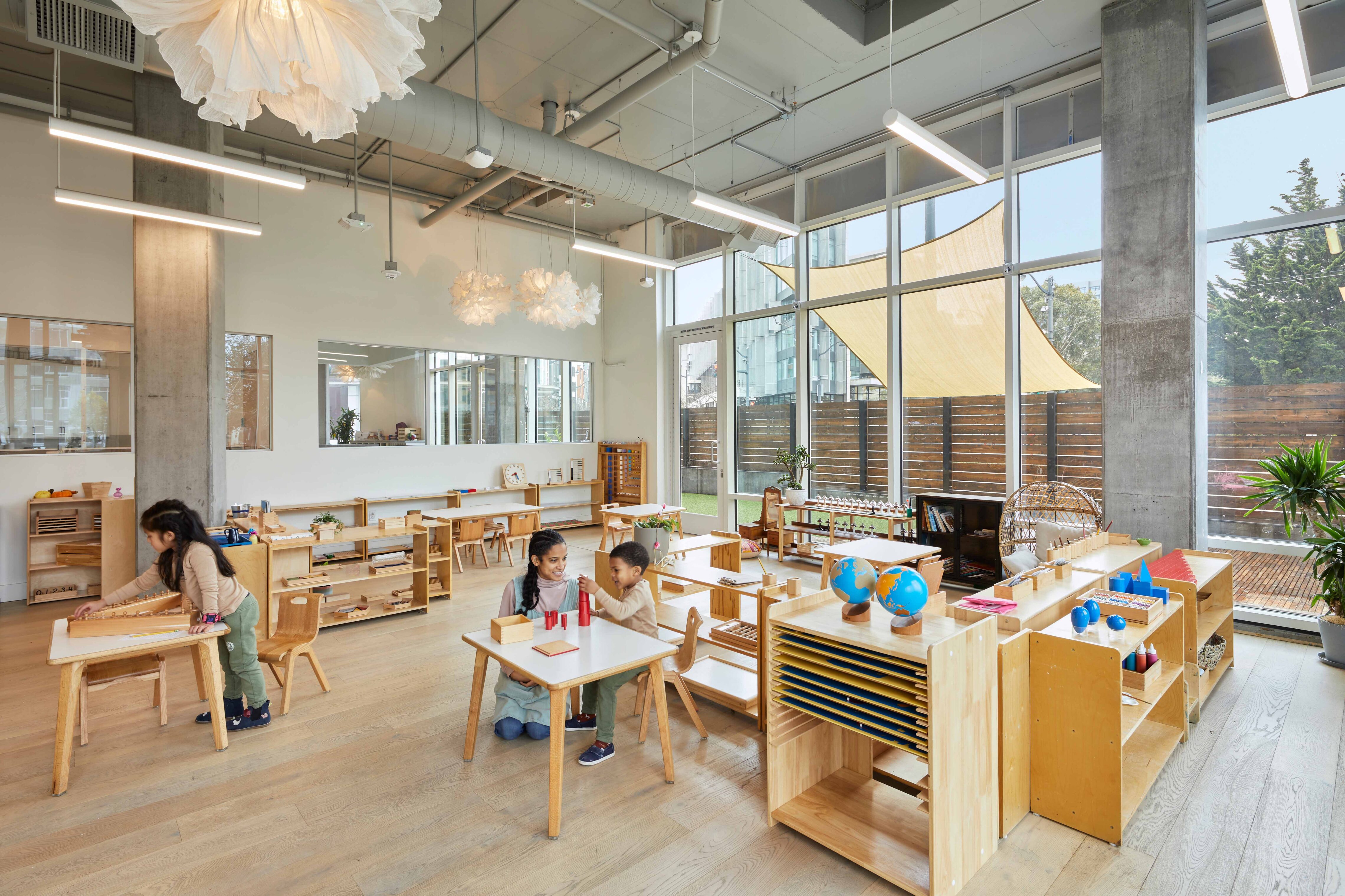 Children working at small wooden tables in an open, light-filled preschool classroom with shelves of learning materials and large windows.