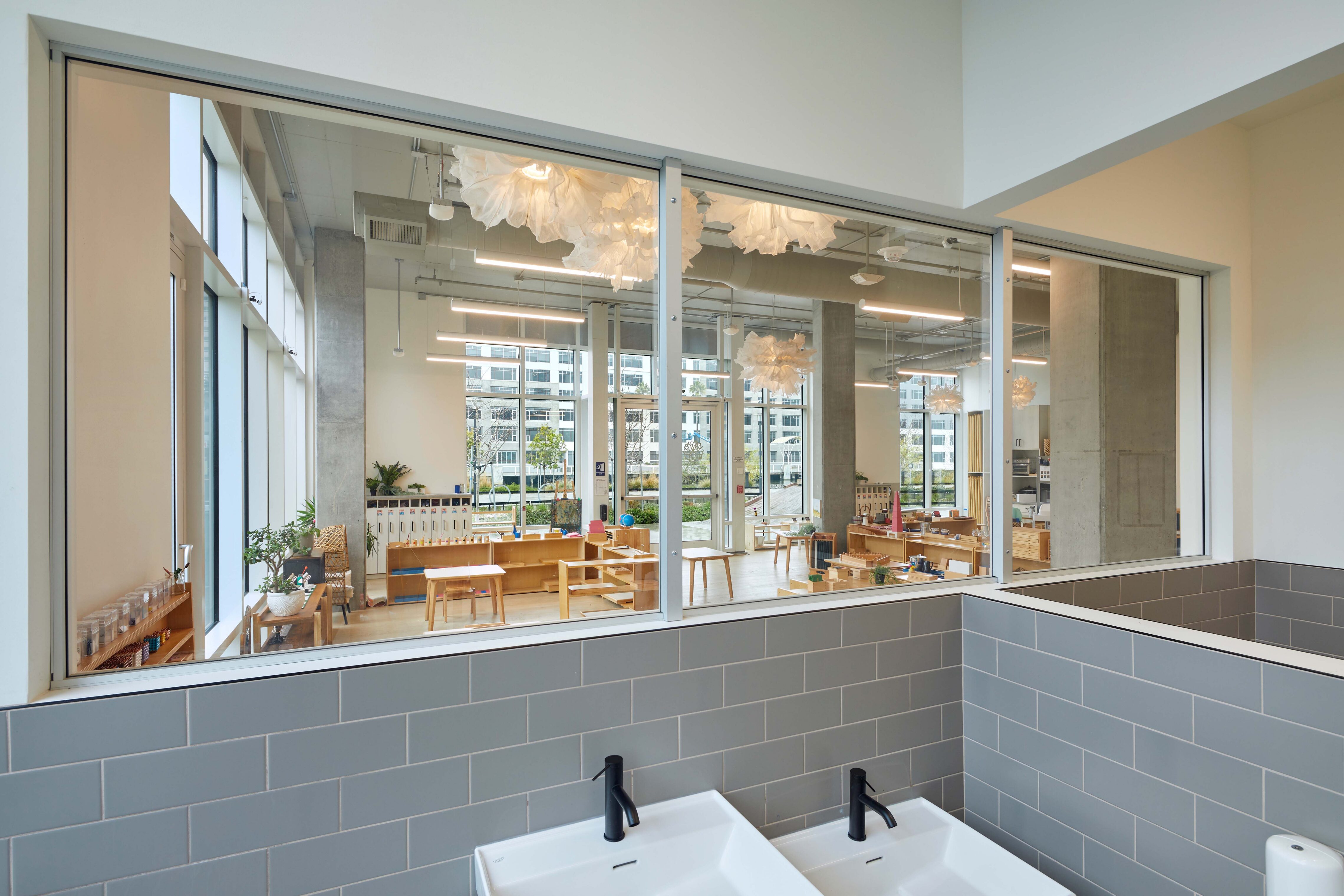 Bathroom sinks in the foreground with a large interior window looking into a bright preschool classroom filled with wooden furniture and learning materials.