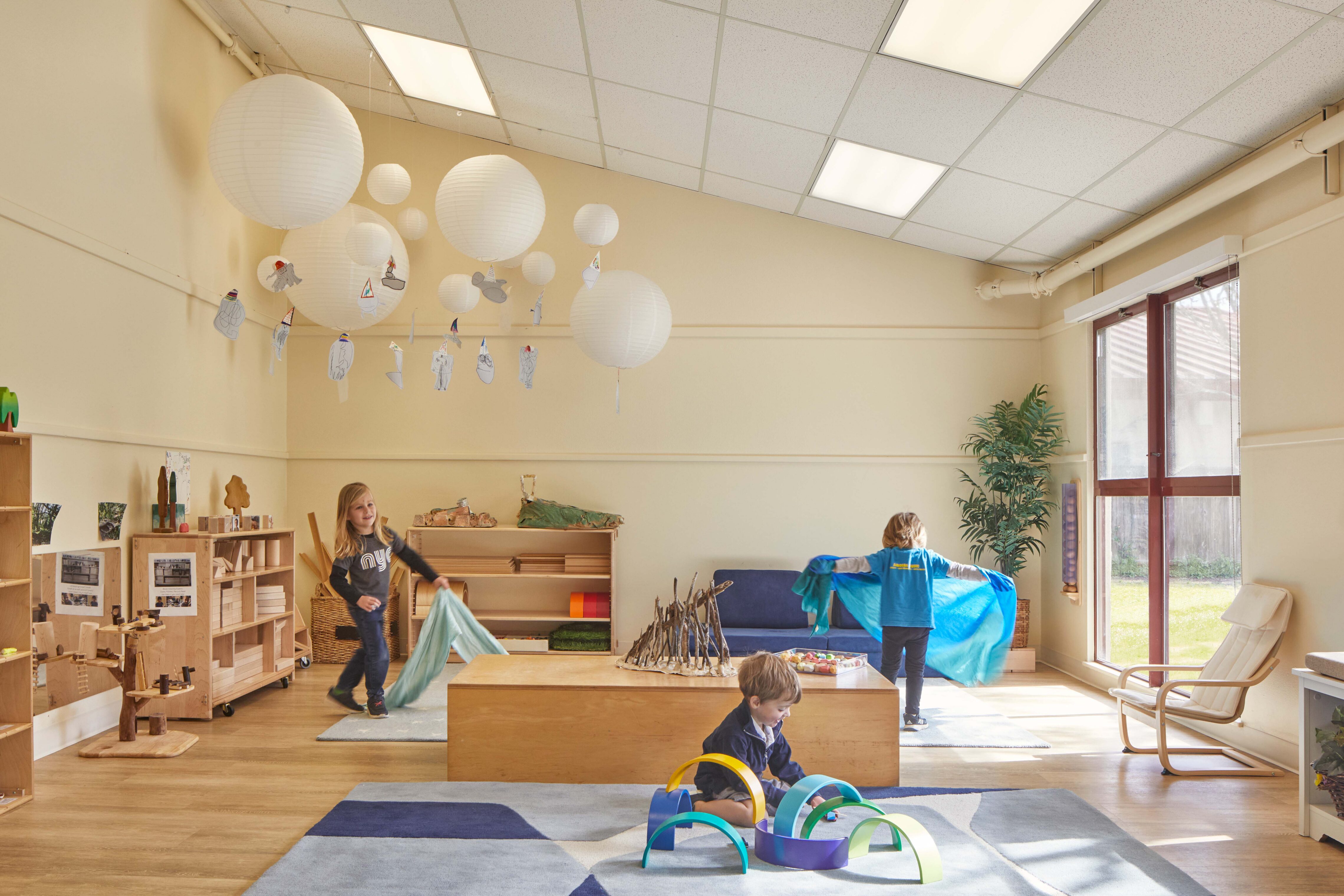 Children play with fabric and building materials in a bright classroom with natural wood furniture and hanging paper lanterns.