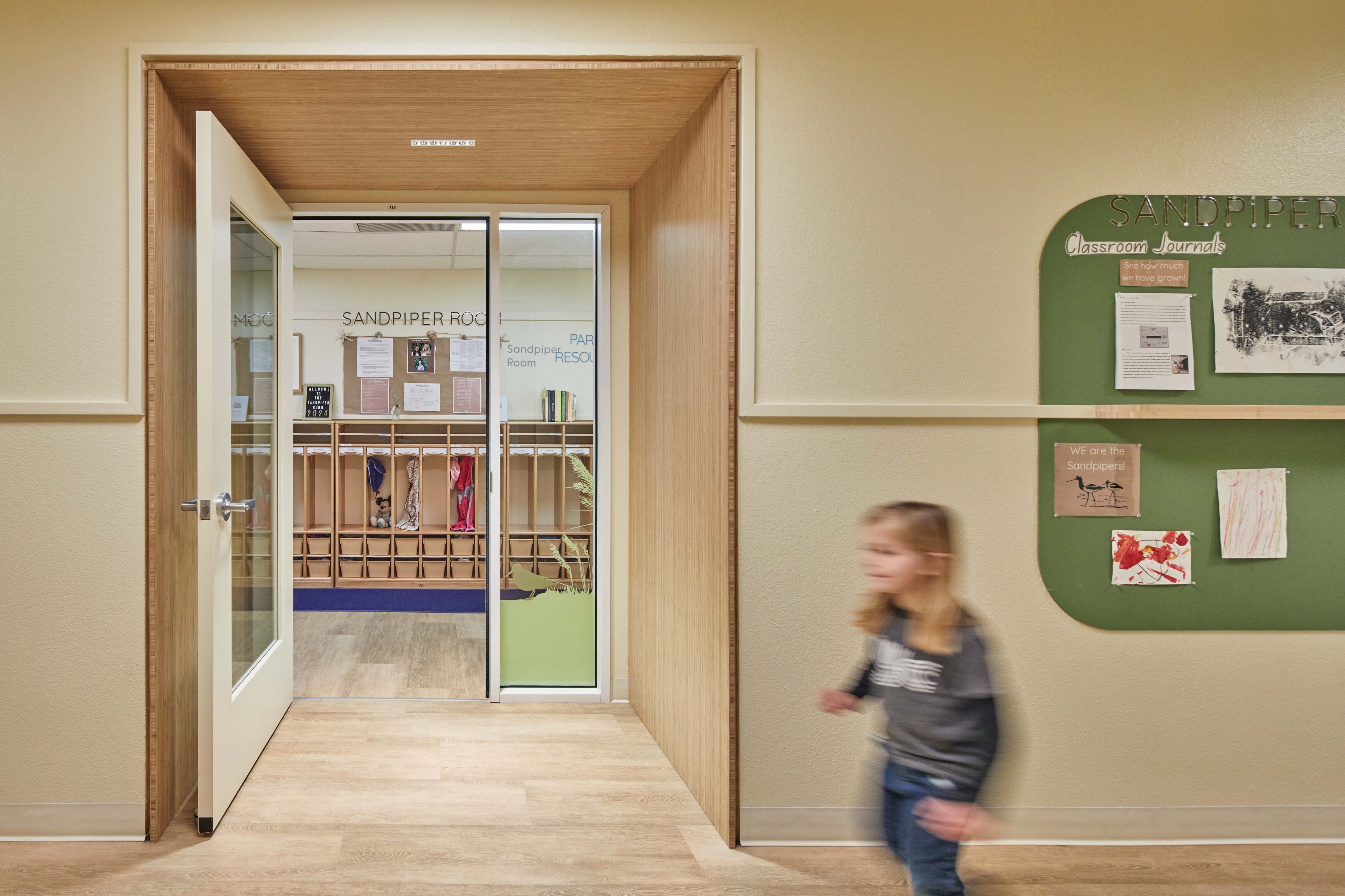 A child walks past an open classroom doorway framed in wood, with student work displayed on a wall-mounted board in the hallway.