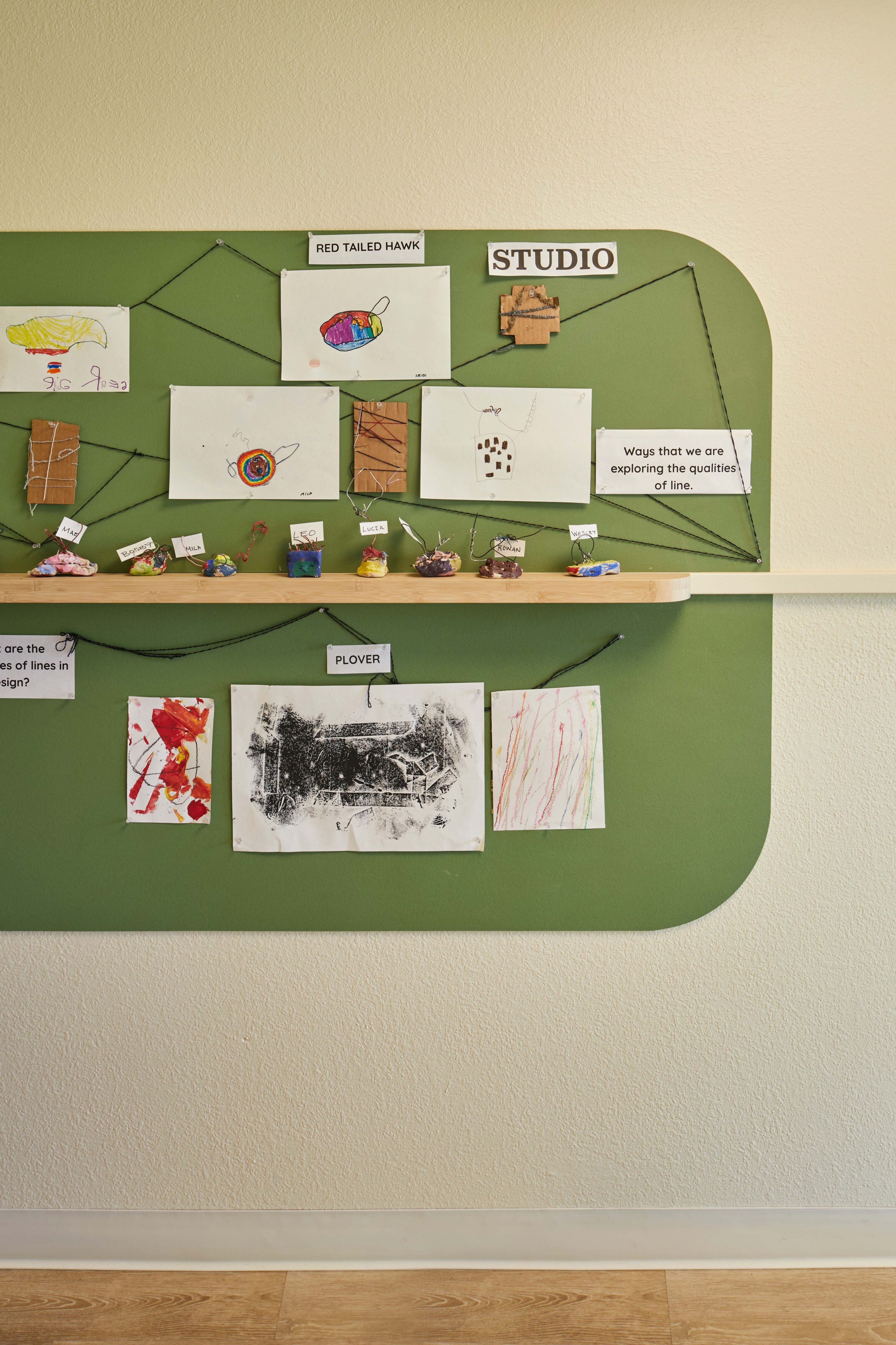 Children’s artwork and small sculptures displayed on a green wall-mounted learning board with labels and string connections.
