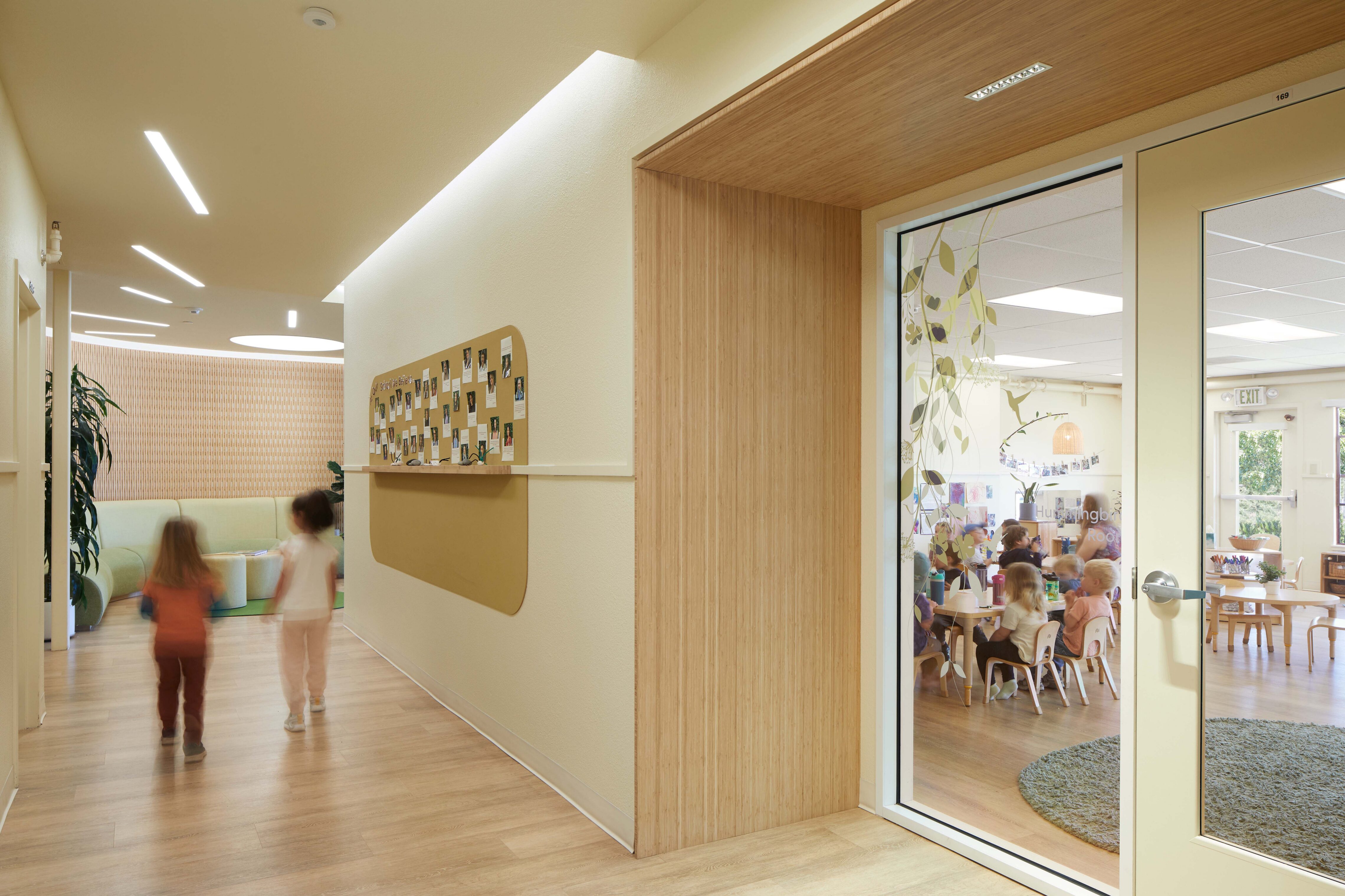 Children walk through a hallway with windows into classrooms and a wall displaying student photos.