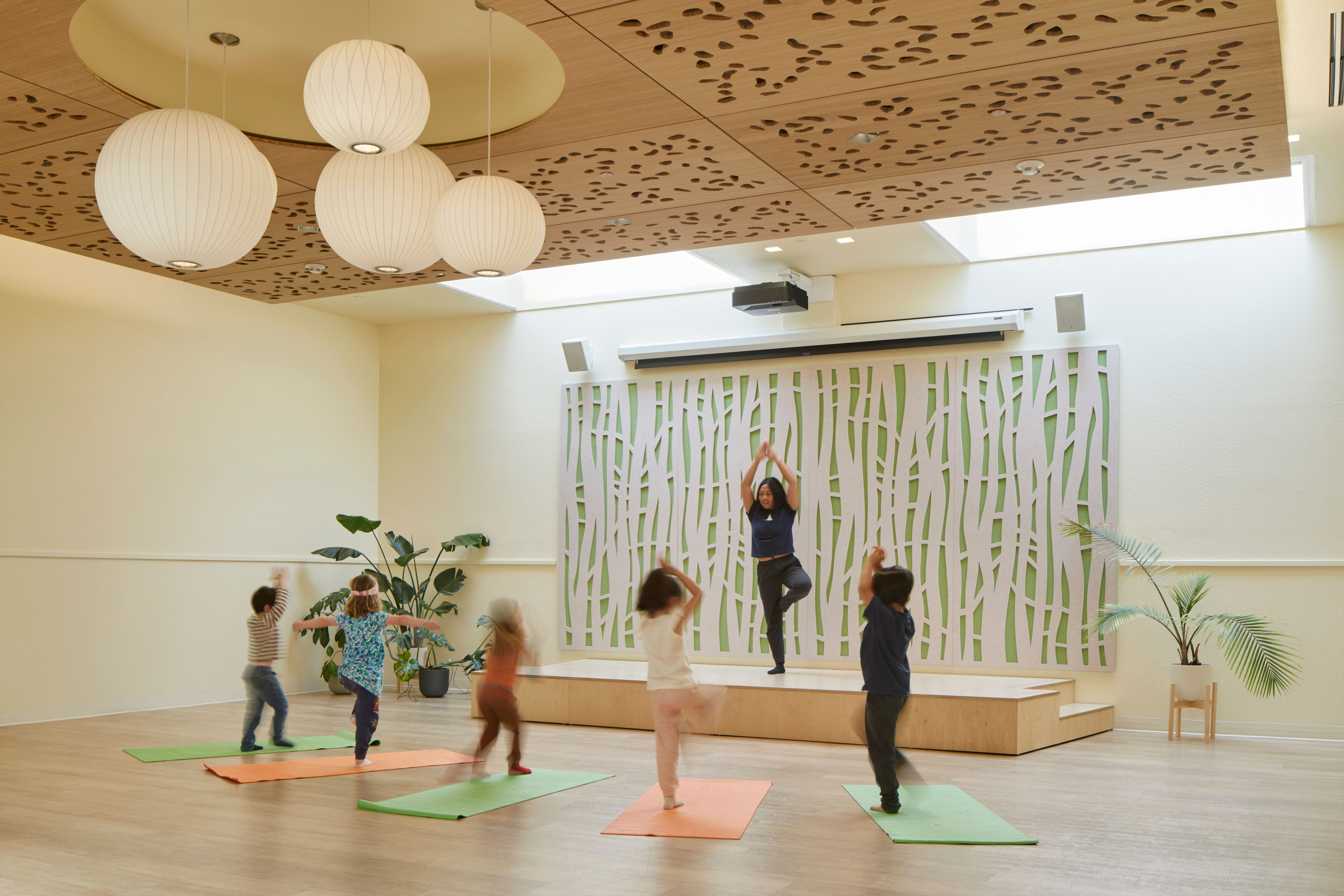 Children work independently at tables in a bright classroom with large windows, open shelving, and orange and green yoga mats.
