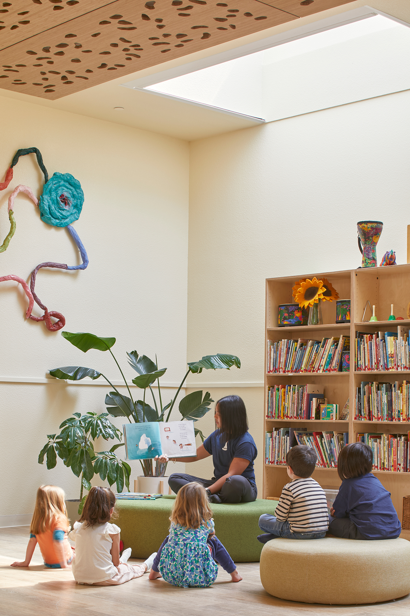 Teacher reads a book to children seated on soft cushions in a cozy library area with tall bookshelves and plants.