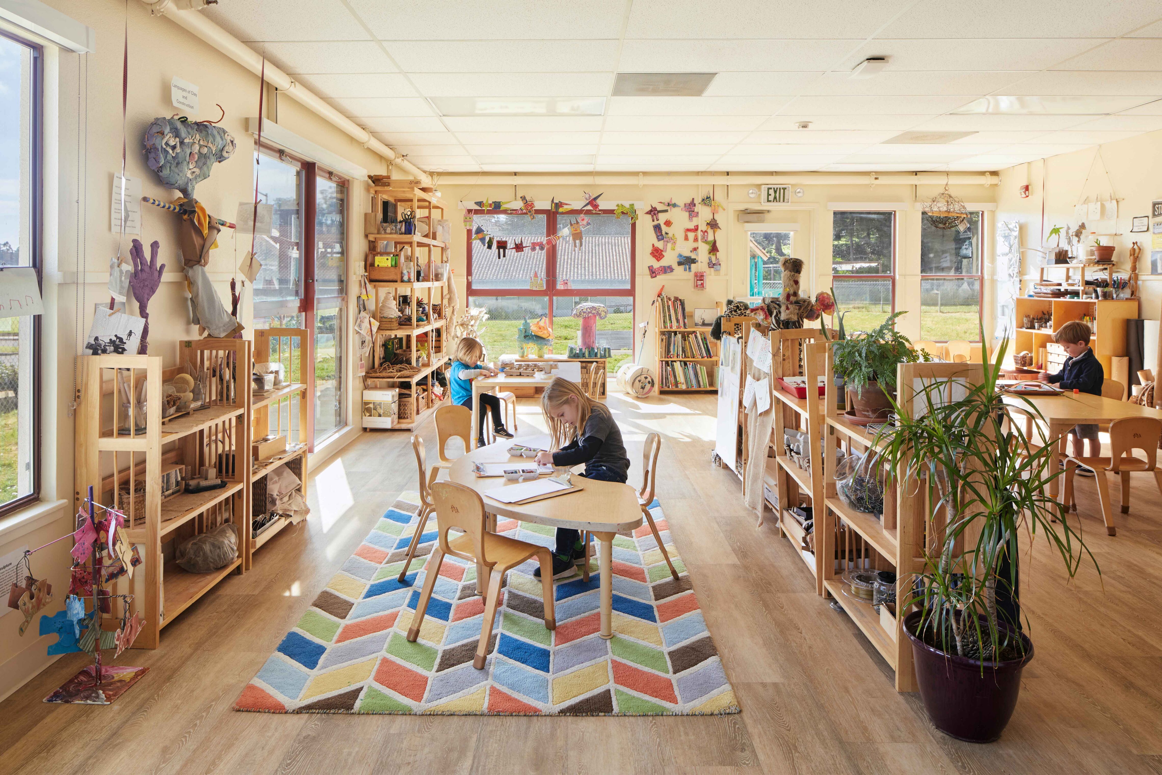 Children work independently at tables in a bright classroom with large windows, open shelving, and colorful rugs.