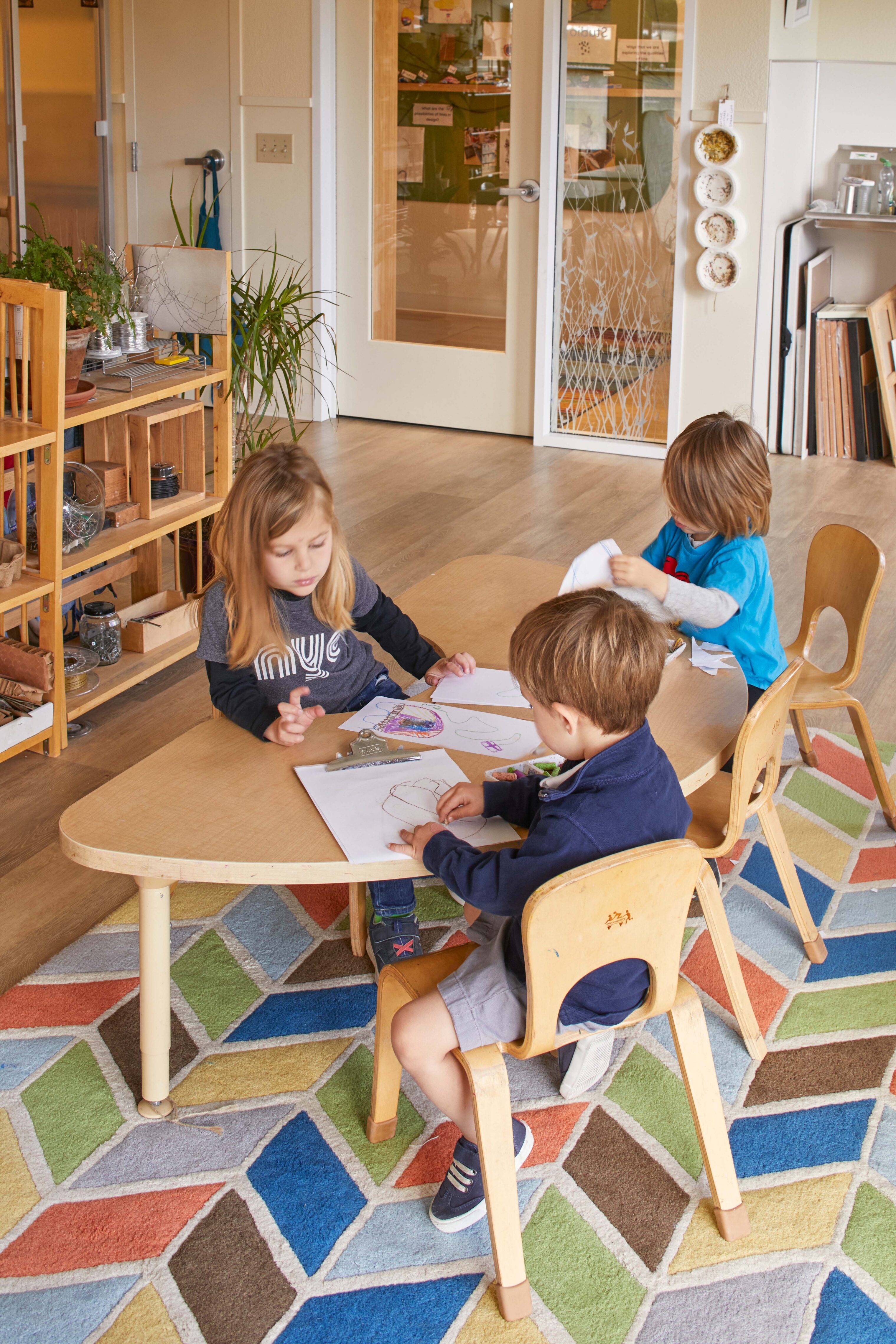 Three young children sit around a low table drawing and working with paper in a classroom filled with natural materials and plants.