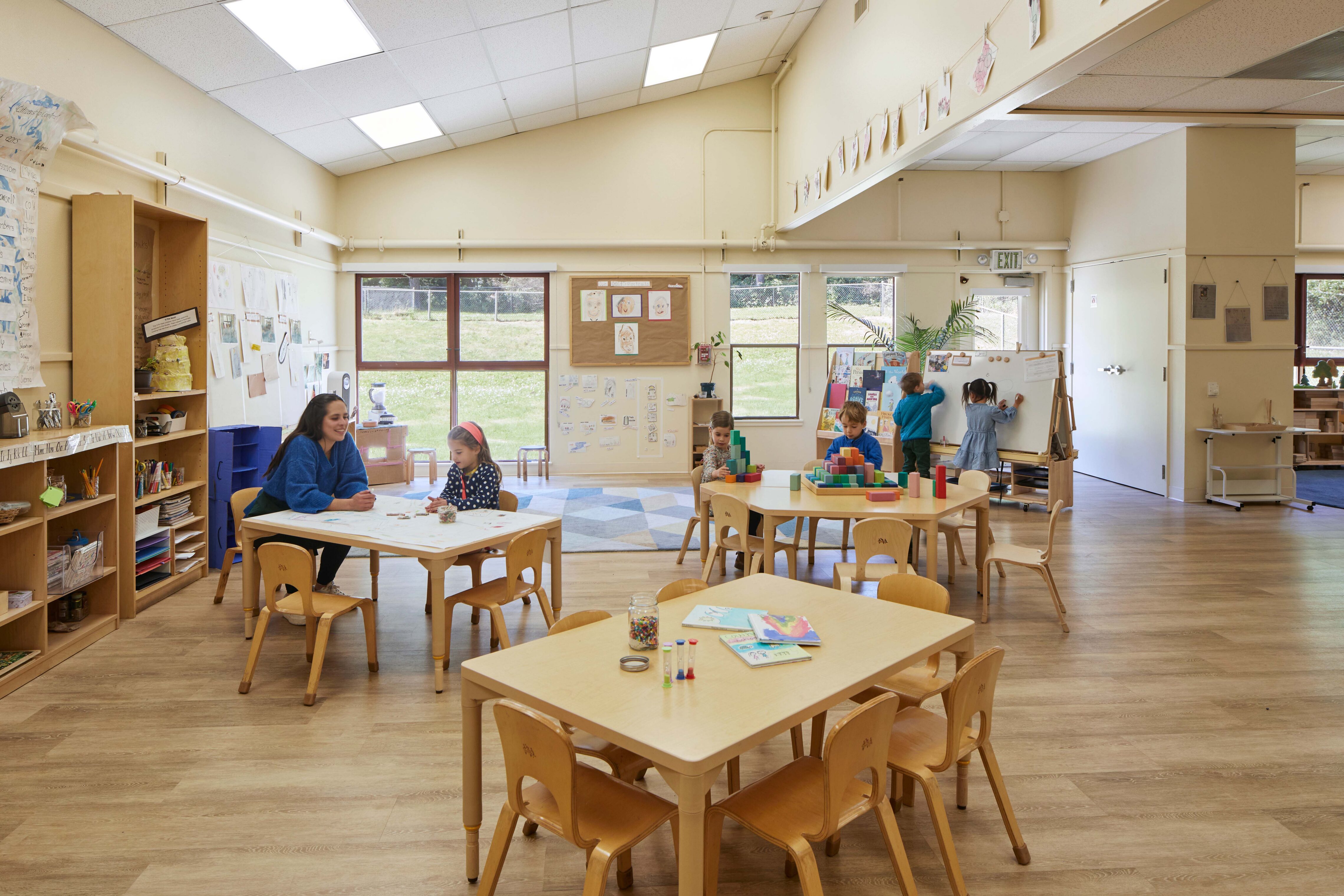 Teacher and children work at small tables with art materials in a spacious classroom with large windows and open activity areas.
