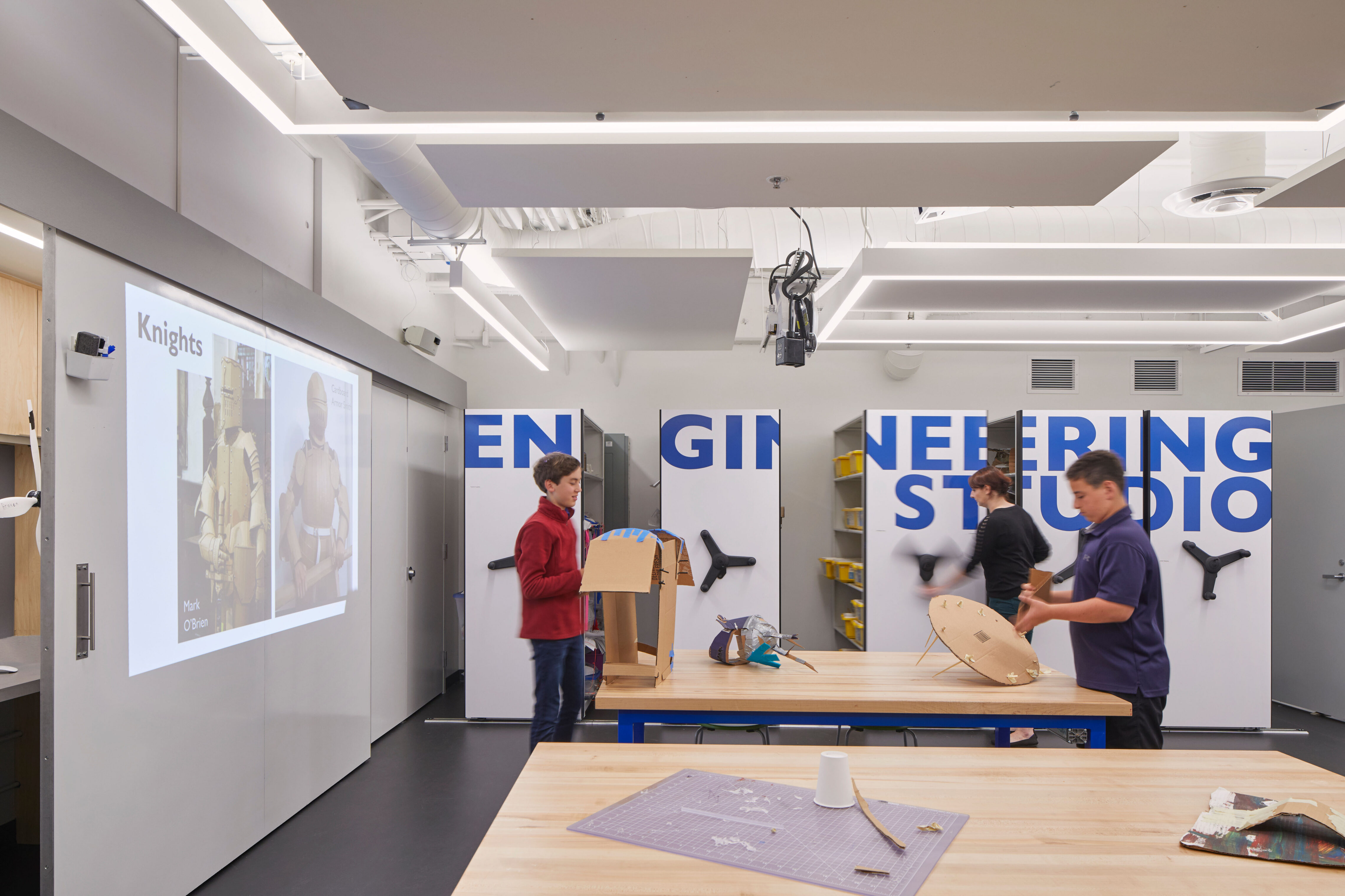 Students test and assemble cardboard prototypes at central worktables beneath suspended power reels and acoustic panels.
