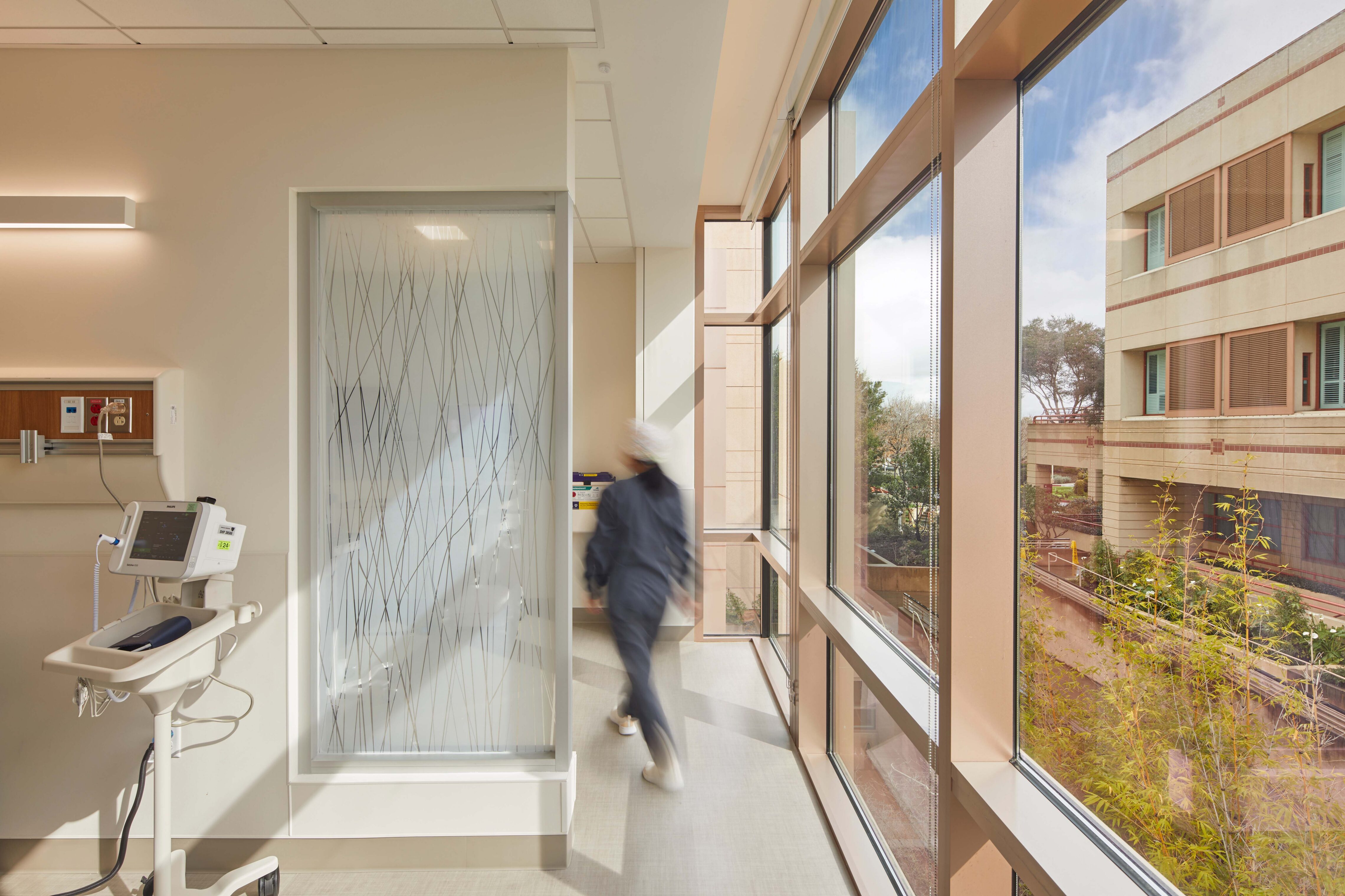 Daylit hospital infusion clinic corridor with a clinician walking past large windows, patterned privacy glass, wall-mounted medical equipment, and exterior courtyard views.