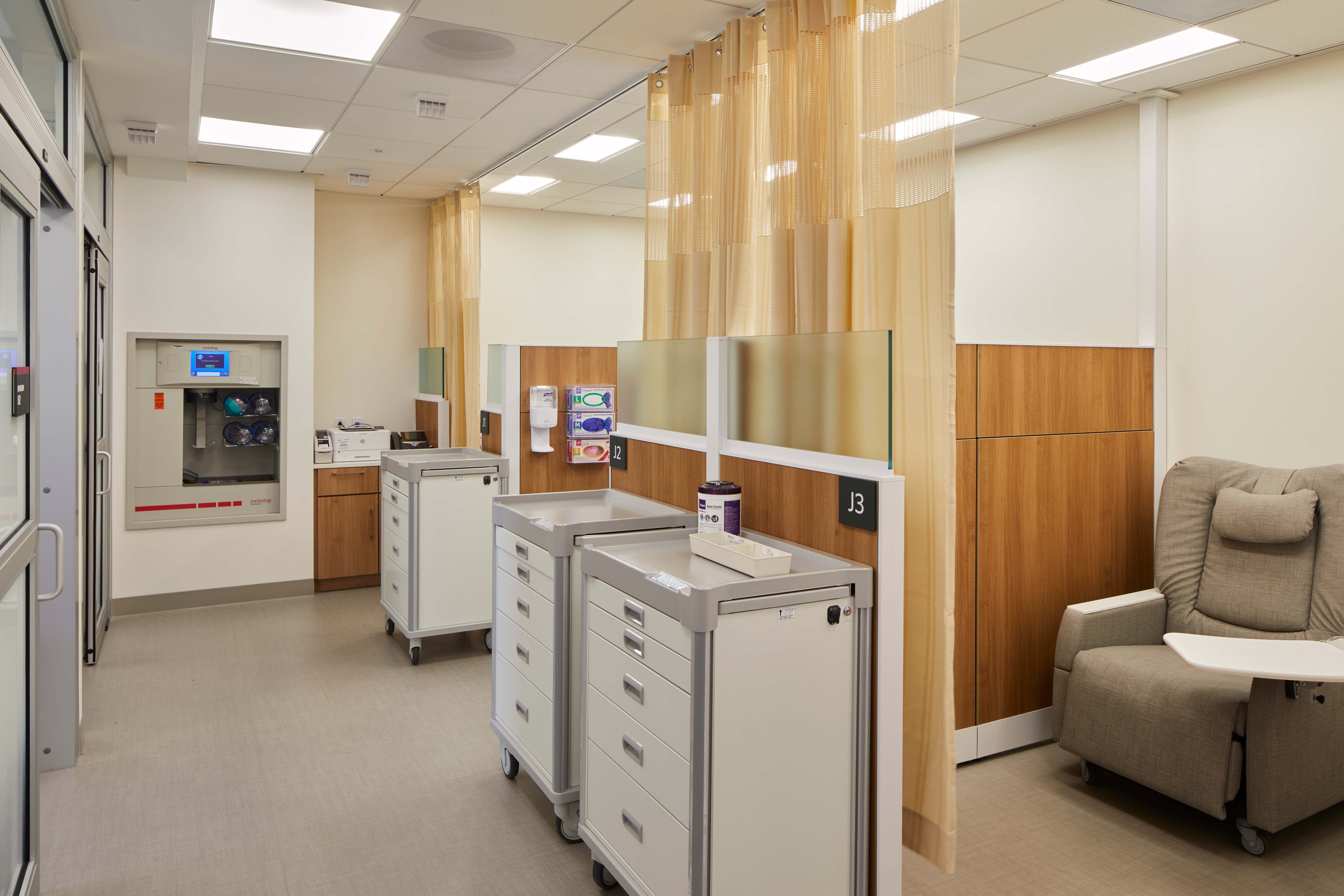 Hospital infusion treatment area with curtained patient bays, recliner chairs, medical carts, supply cabinets, and warm wood wall panels.
