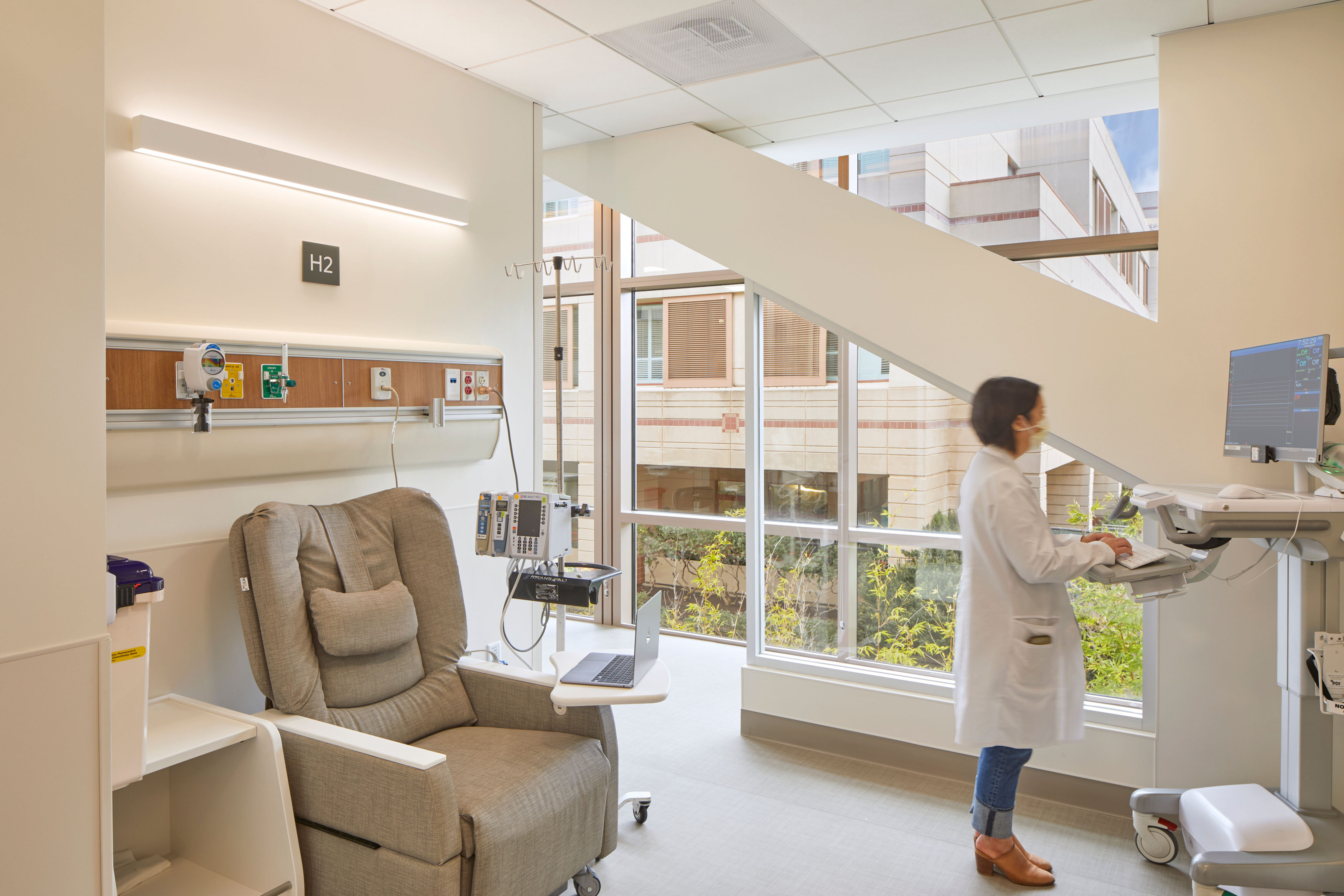 Daylit hospital infusion treatment room with a recliner, wall-mounted medical equipment, large windows, courtyard views, and a clinician working at a patient monitor.