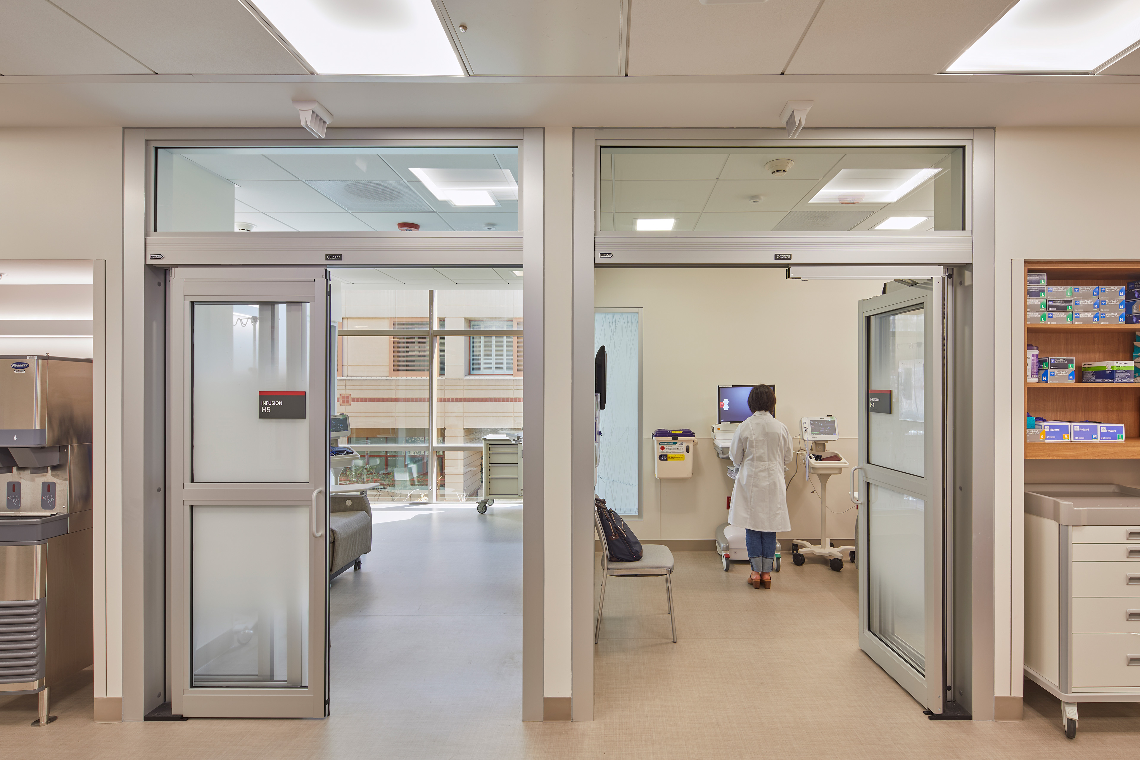 Infusion treatment area with two glass-fronted patient bays, a clinician reviewing equipment, integrated medical storage, and daylight from large interior-facing windows.