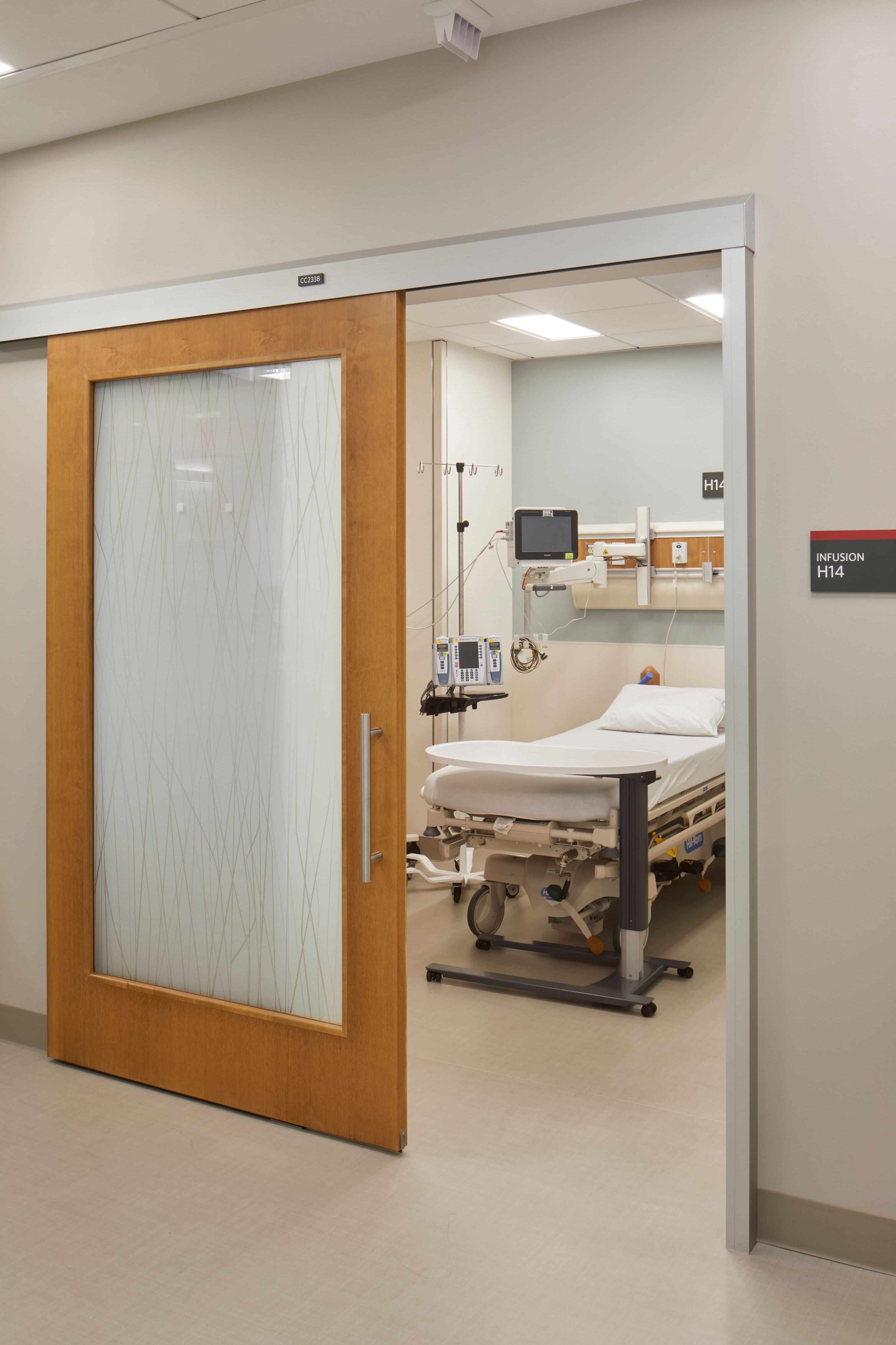 Private infusion treatment room with a sliding wood door, patterned privacy glass, patient bed, bedside table, and medical monitoring equipment.
