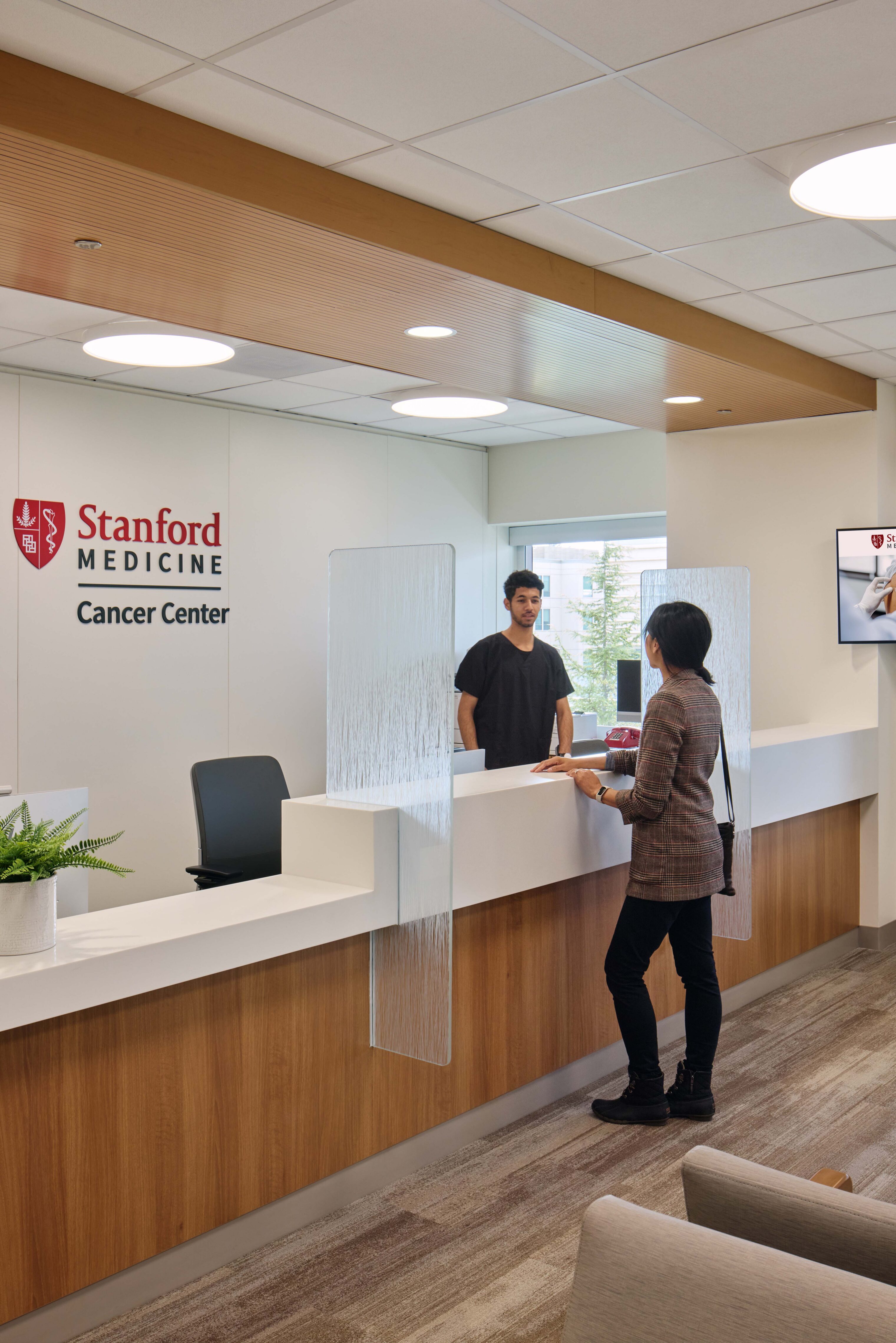 Stanford Medicine Cancer Center reception desk with wood finishes, textured glass dividers, staff member, and patient checking in.