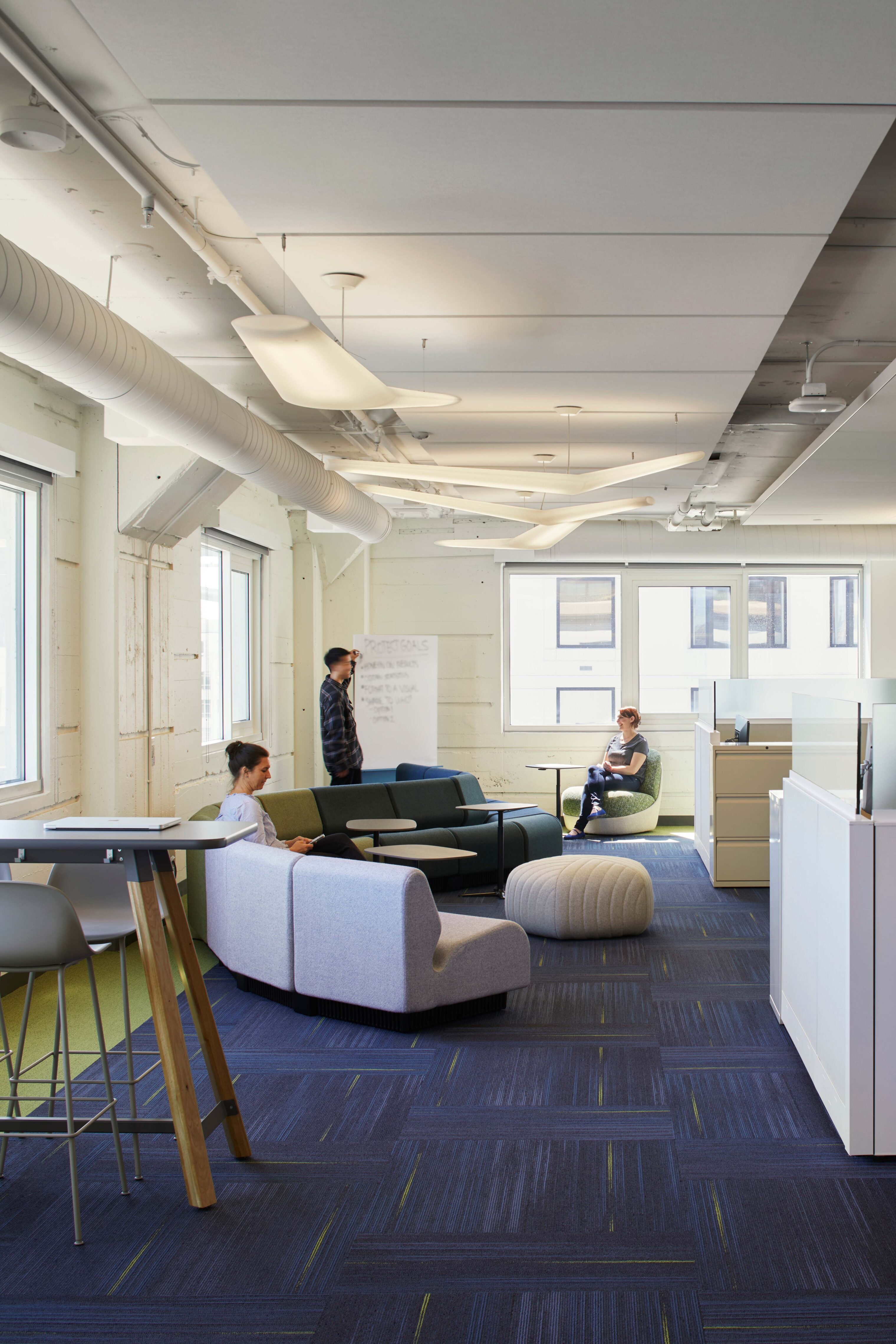 Open office lounge with soft seating, high tables, workstations, and sculptural pendant lights, supporting both focused work and informal collaboration.