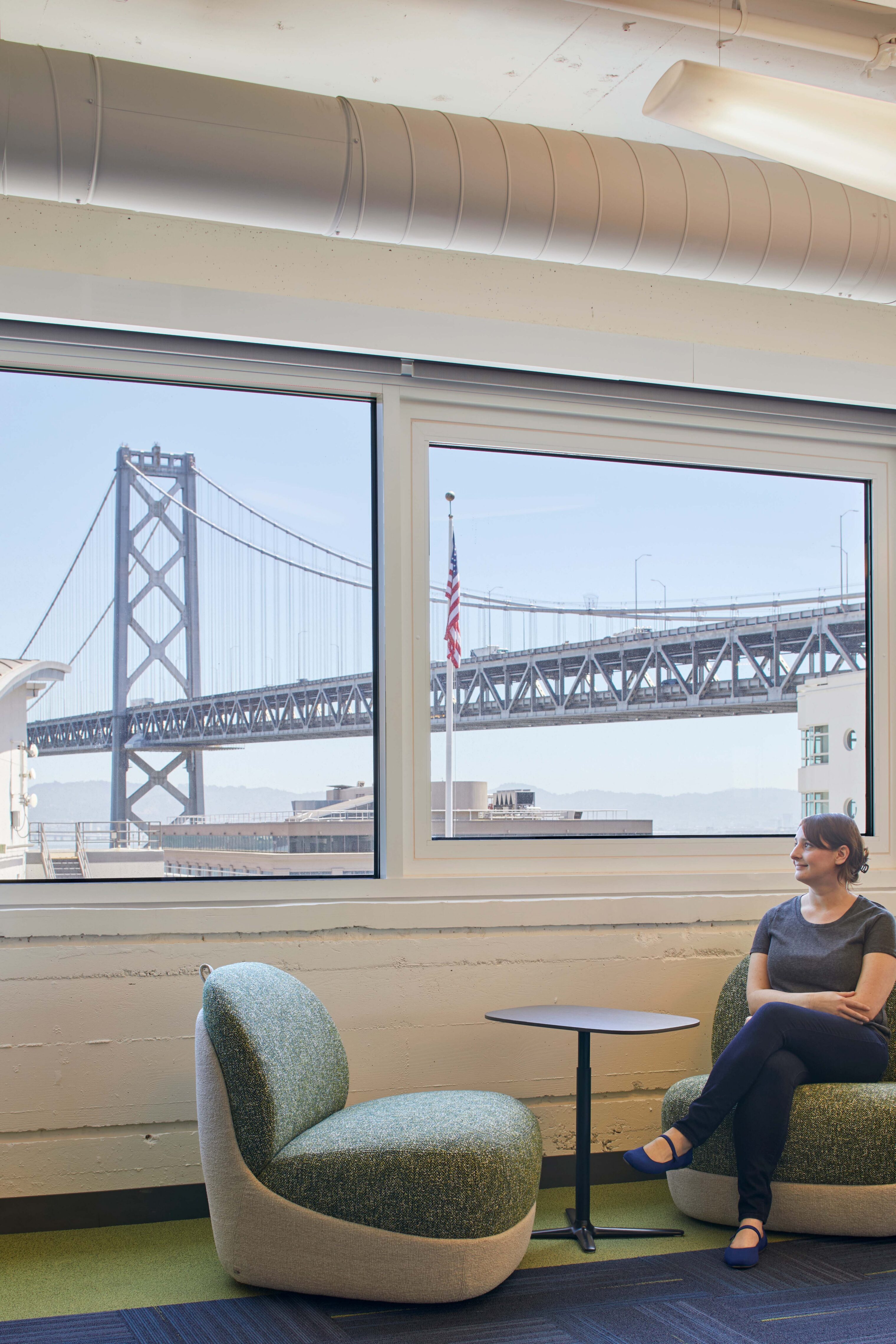 Workplace lounge with soft seating, exposed ceiling systems, and large windows framing views of the Bay Bridge.
