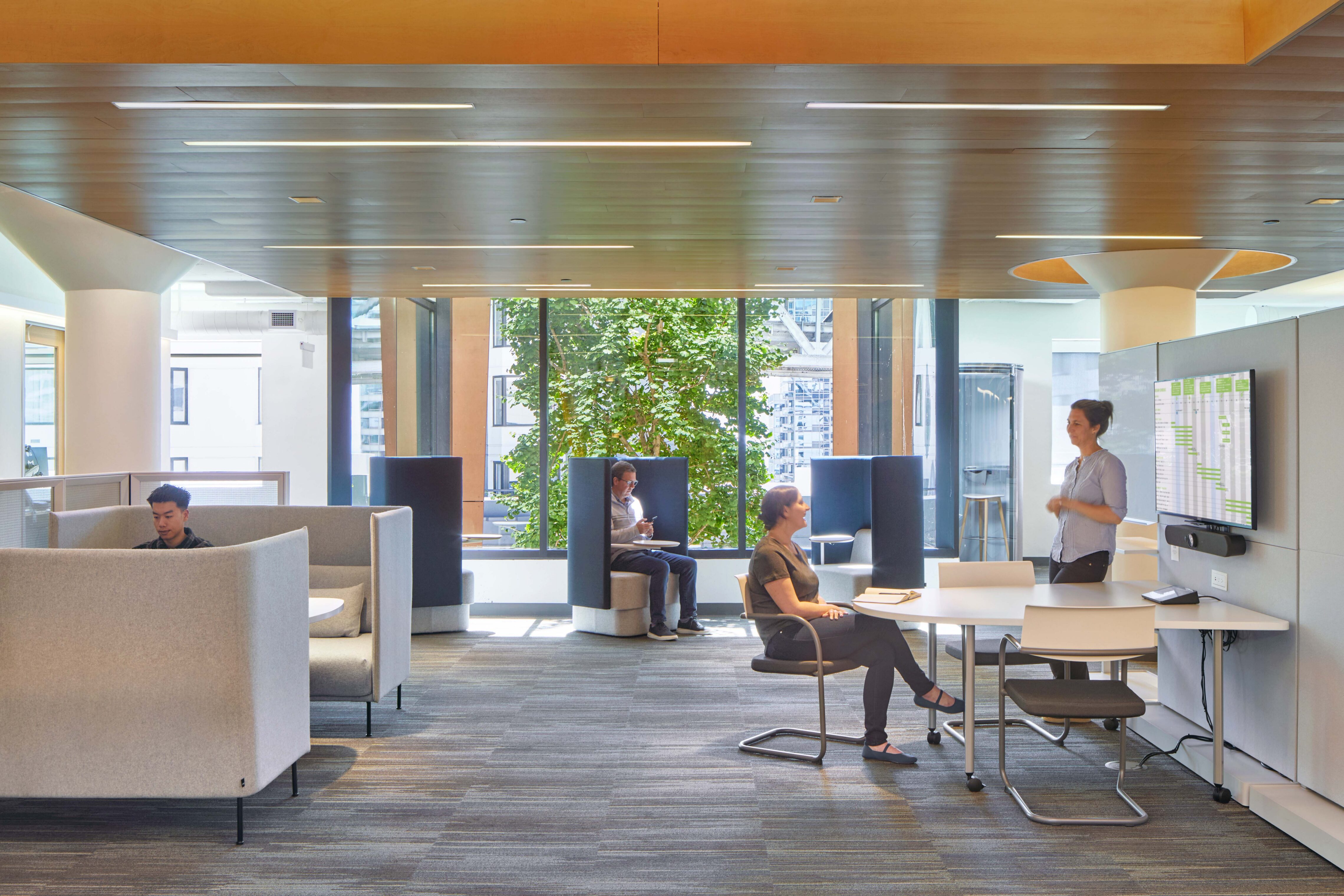 Bright workplace collaboration area with high-back lounge seating, open workstations, a presentation screen, and large windows overlooking the city.