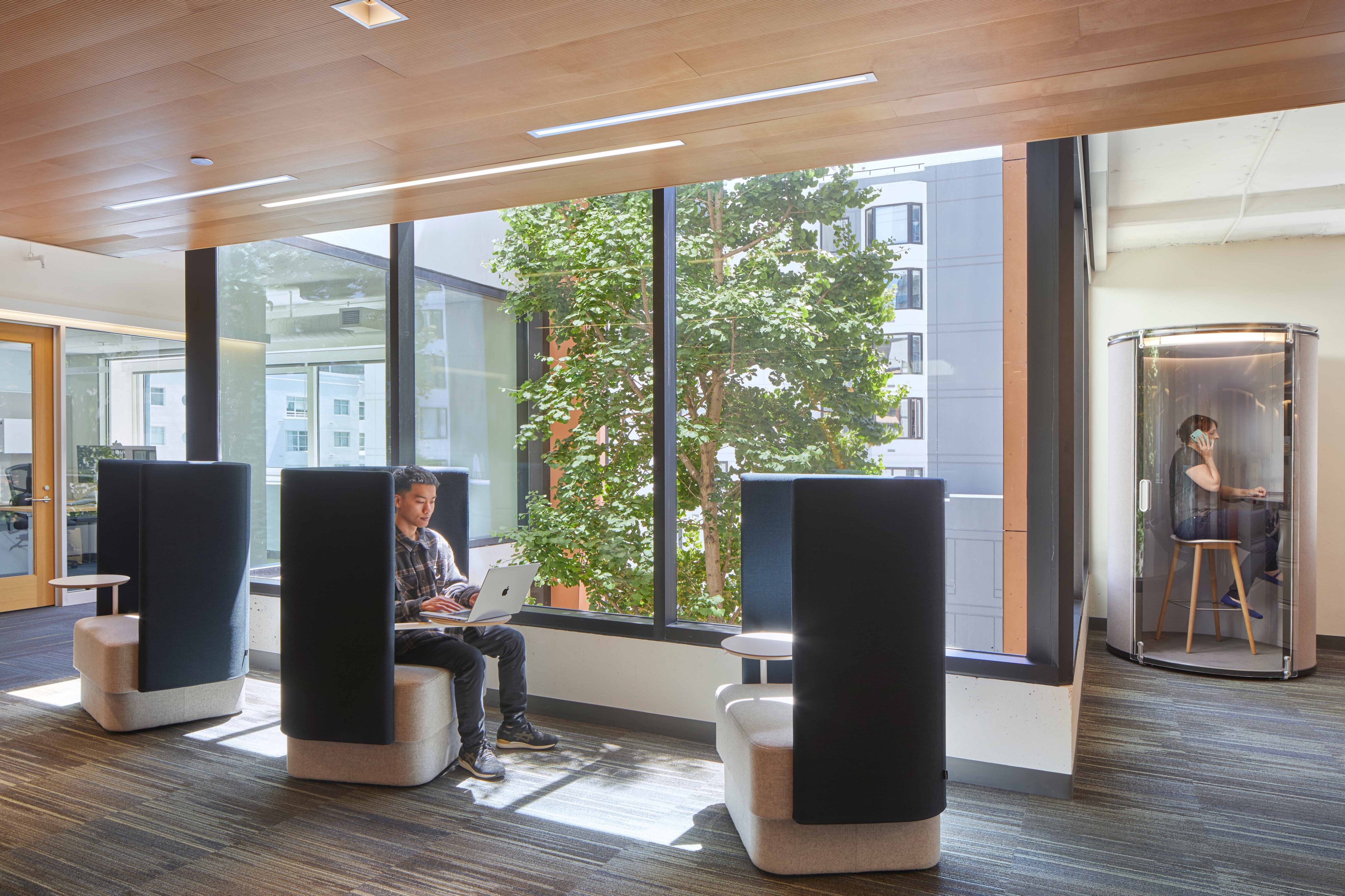 Open workplace lounge with high-back privacy seating, large windows, and an enclosed phone booth, supporting both focused individual work and private calls.
