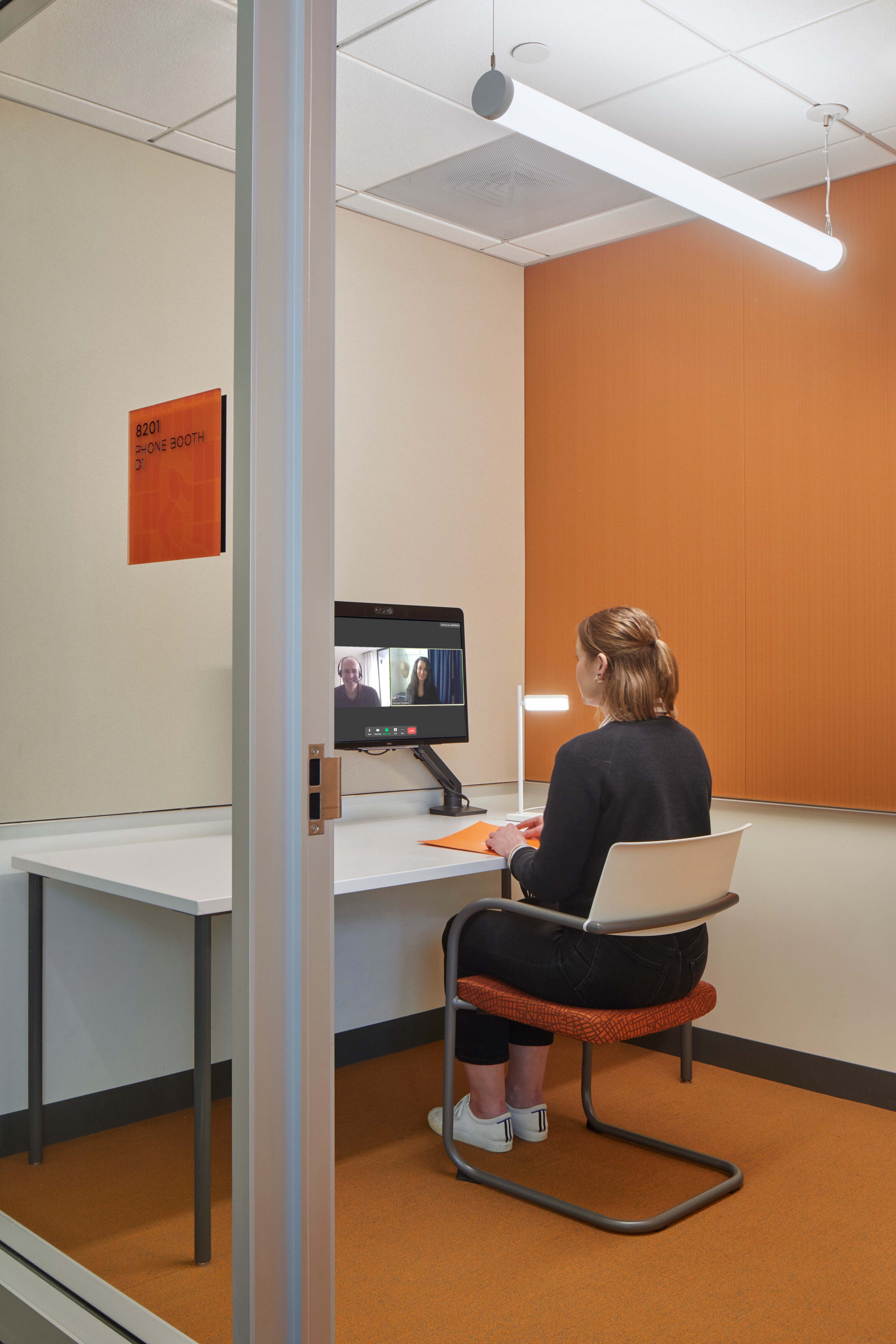 Private phone room with an orange accent wall, desk, task lighting, and video conferencing setup for focused individual calls.