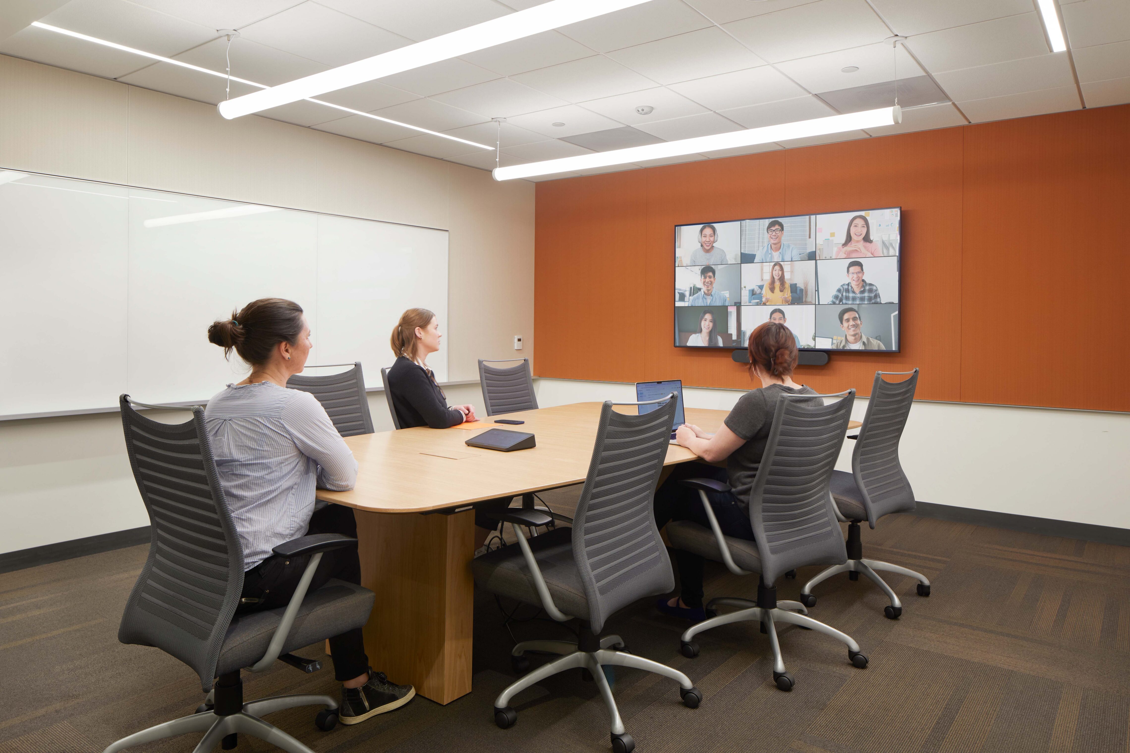 Conference room with a large meeting table, whiteboard wall, and video conferencing display connecting in-person participants with remote attendees.