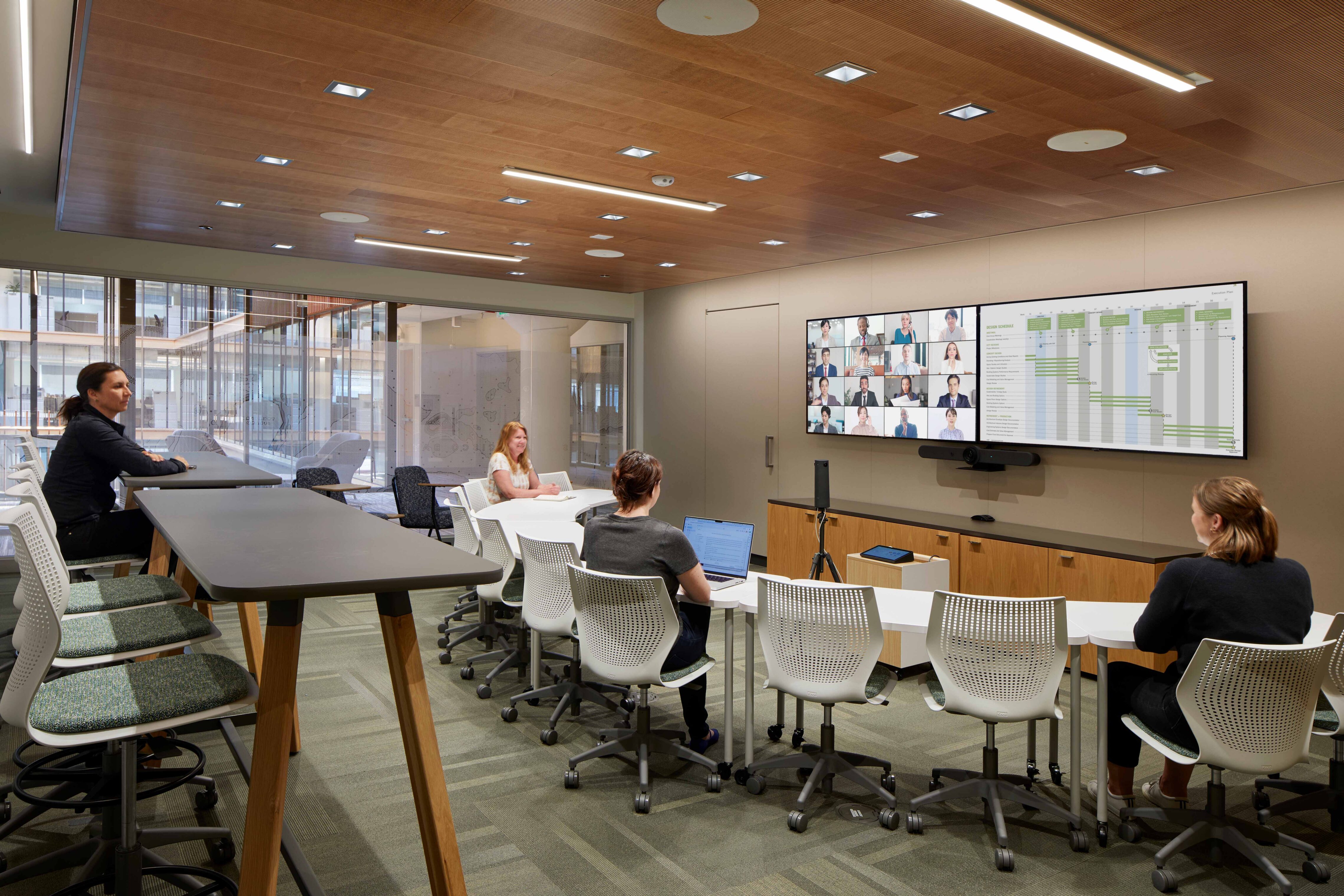 A meeting room with flexible seating, high tables, wood ceiling accents, and integrated video conferencing technology supporting in-person and remote collaboration.